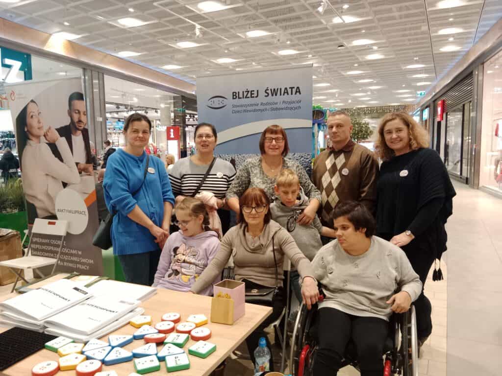 Grupa dorosłych i dzieci, w tym jedno na wózku inwalidzkim, pozuje przy stoliku z materiałami edukacyjnymi i banerem Bliżej Świata w centrum handlowym.