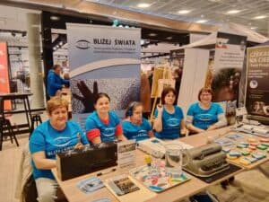 Pięć kobiet w niebieskich koszulkach "Bliżej Świata" siedzi przy stoisku z materiałami informacyjnymi i rękodziełem na imprezie wewnętrznej lub targach.