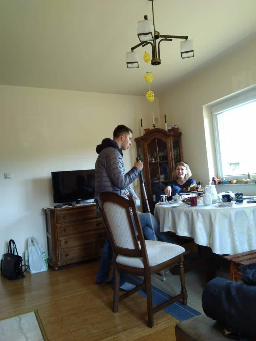 Mężczyzna stoi i rozmawia z siedzącą kobietą przy stole zastawionym naczyniami w salonie z oknem, telewizorem i drewnianymi meblami.