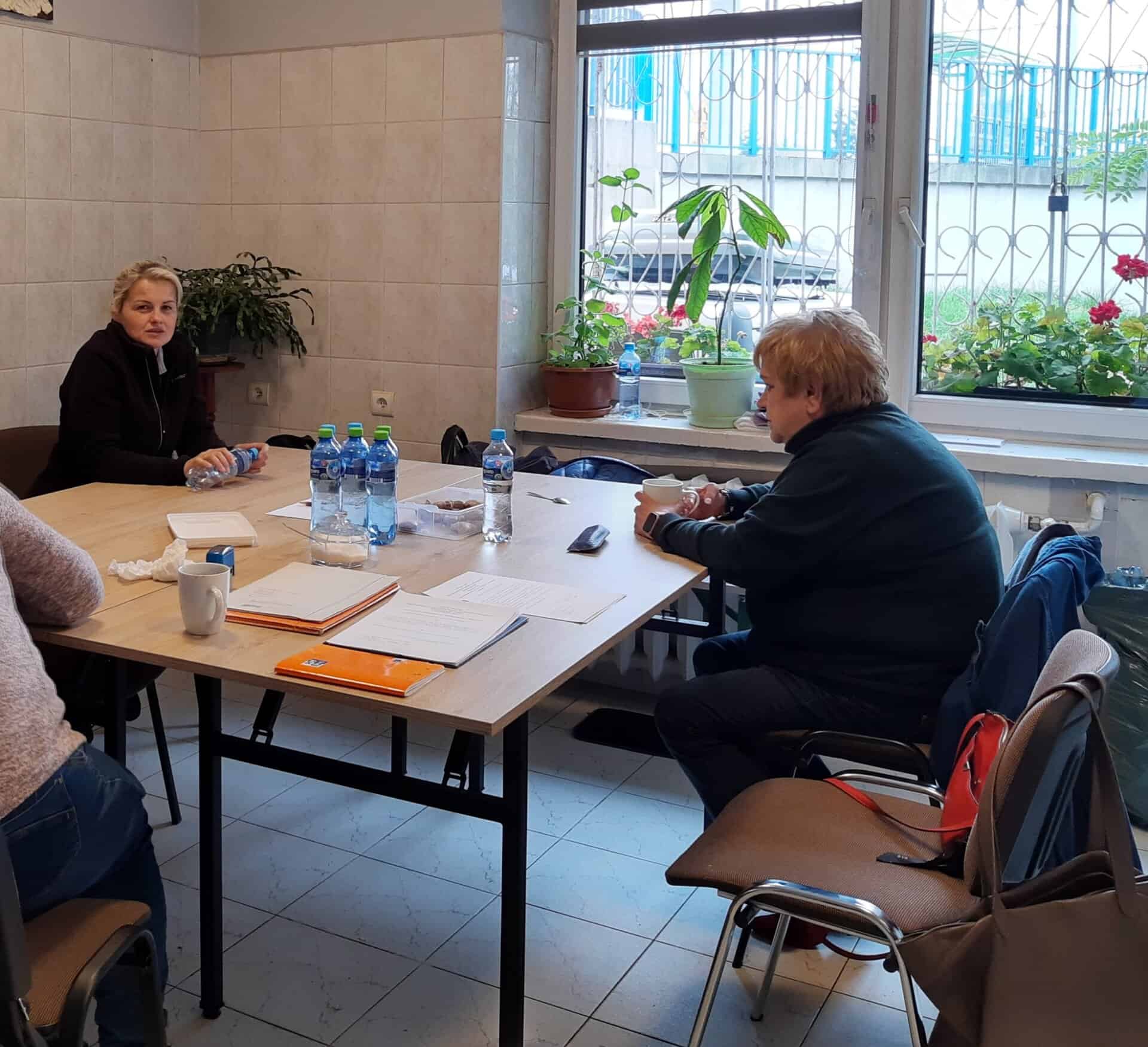 Dwie osoby siedzą przy stole w pokoju z dużymi oknami, butelkami wody, papierami i roślinami na parapecie. Inna osoba jest częściowo widoczna na pierwszym planie.