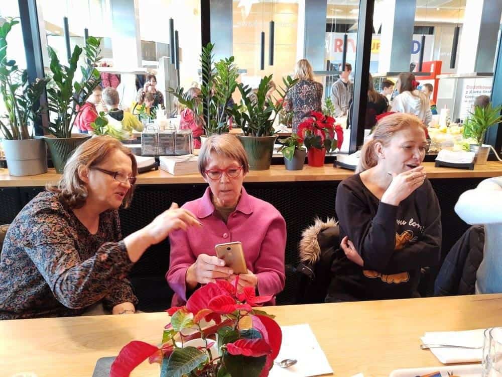 Trzy kobiety siedzą przy stole z roślinami, jedna pokazuje telefon drugiej, podczas gdy trzecia stoi obok i je. W tle widoczni są ludzie i zieleń.