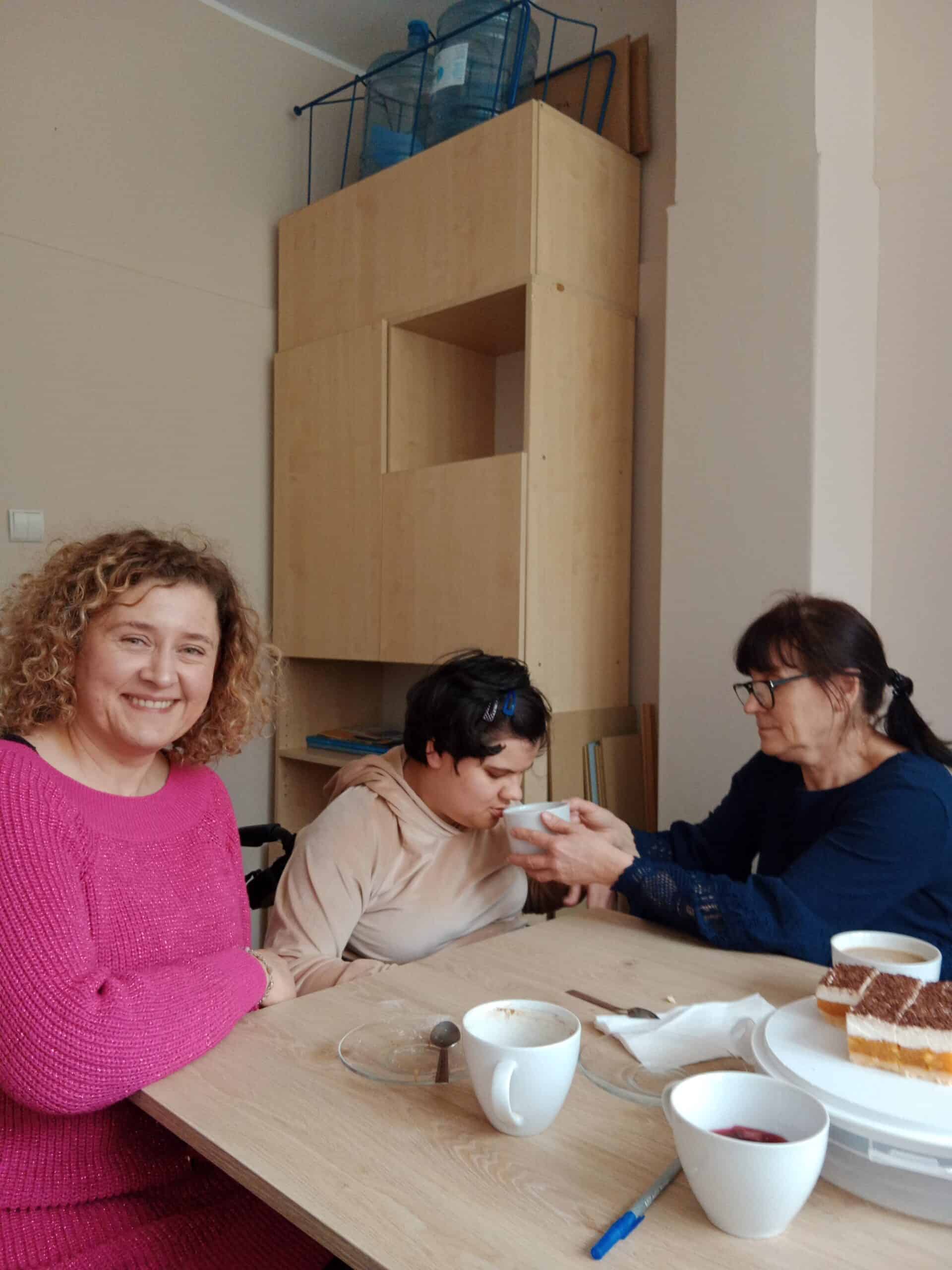 Trzy kobiety siedzą przy stole z kawą i ciastem; jedna pomaga drugiej pić z filiżanki, podczas gdy trzecia uśmiecha się do kamery.