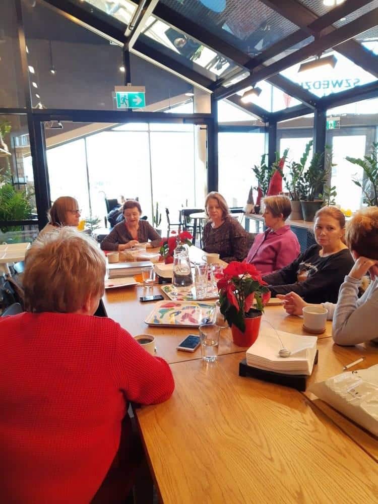 Grupa kobiet siedzi przy drewnianym stole w jasnej kawiarni i prowadzi dyskusję. Na stole leżą papiery, napoje i rośliny doniczkowe.