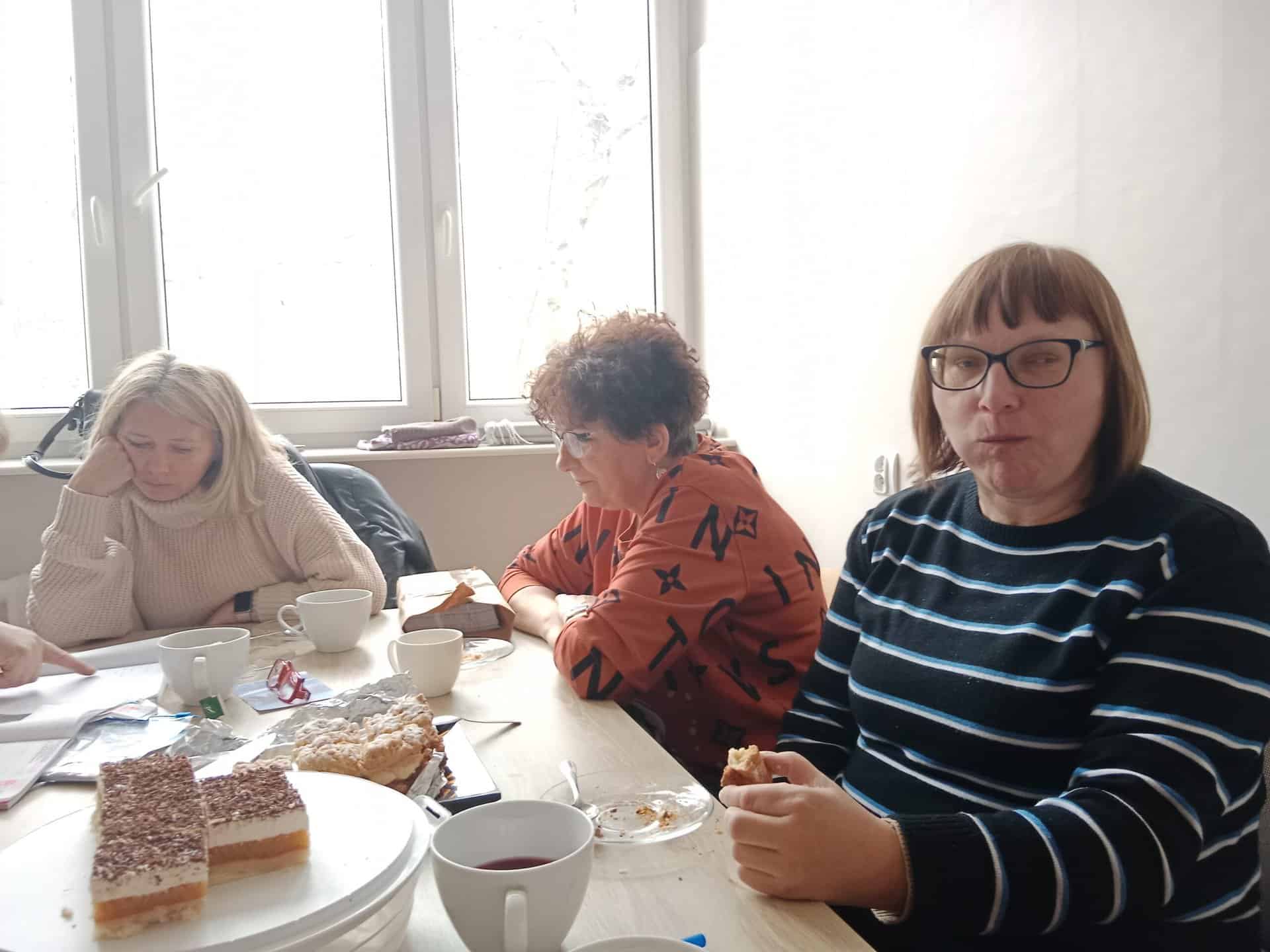 Trzy kobiety siedzą przy stole z kubkami i talerzami ciasta przed sobą, w pobliżu okna, przez które wpada światło dzienne.