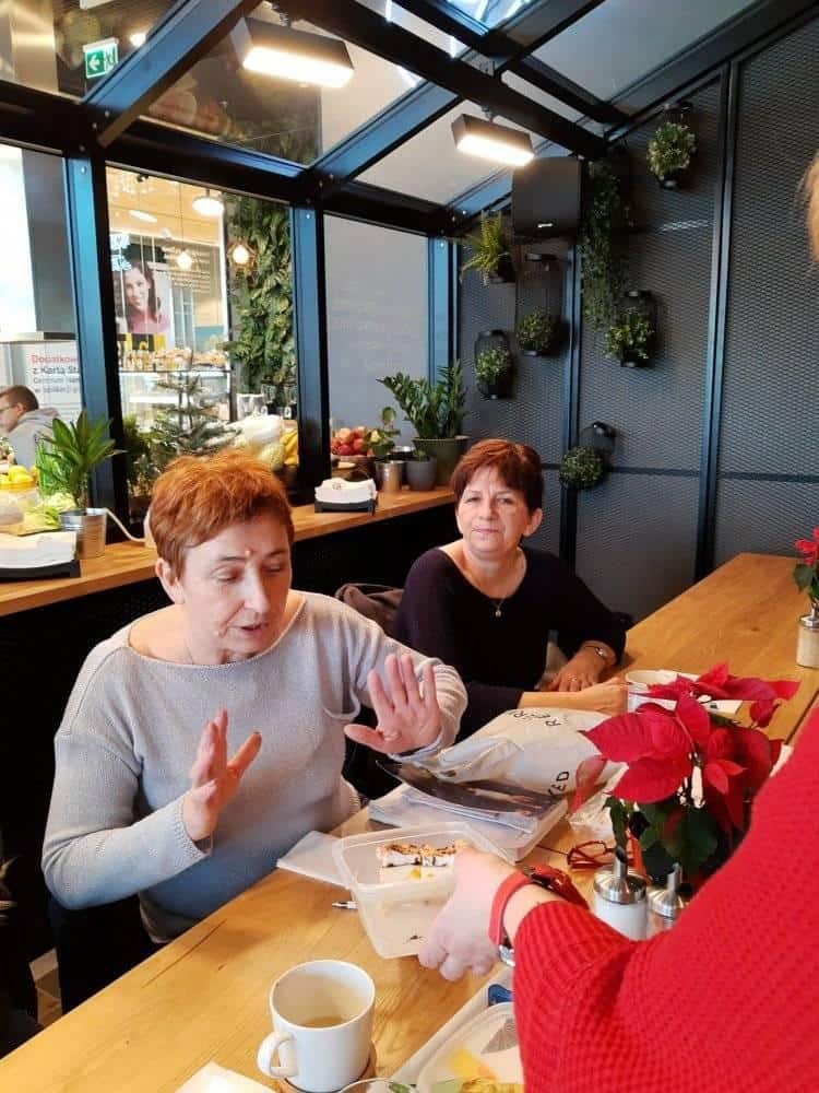 Dwie kobiety siedzą przy stoliku w kawiarni z jedzeniem i napojami; jedna z nich gestykuluje rękami, a druga oferuje coś po drugiej stronie stołu.