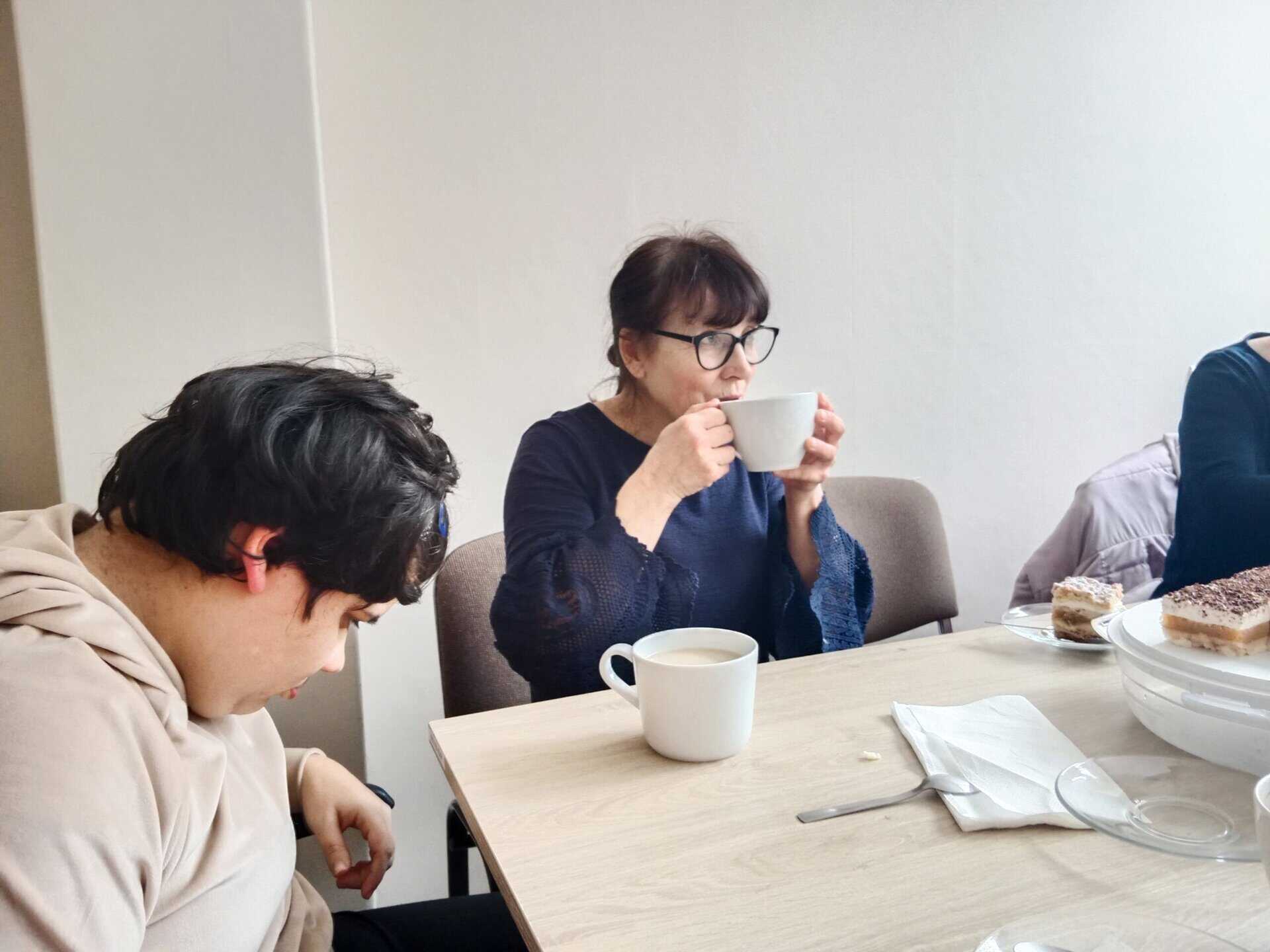 Dwie osoby siedzą przy stole z kawą i ciastem; jedna pije z filiżanki, druga patrzy w dół, a część trzeciej osoby jest widoczna po prawej stronie.
