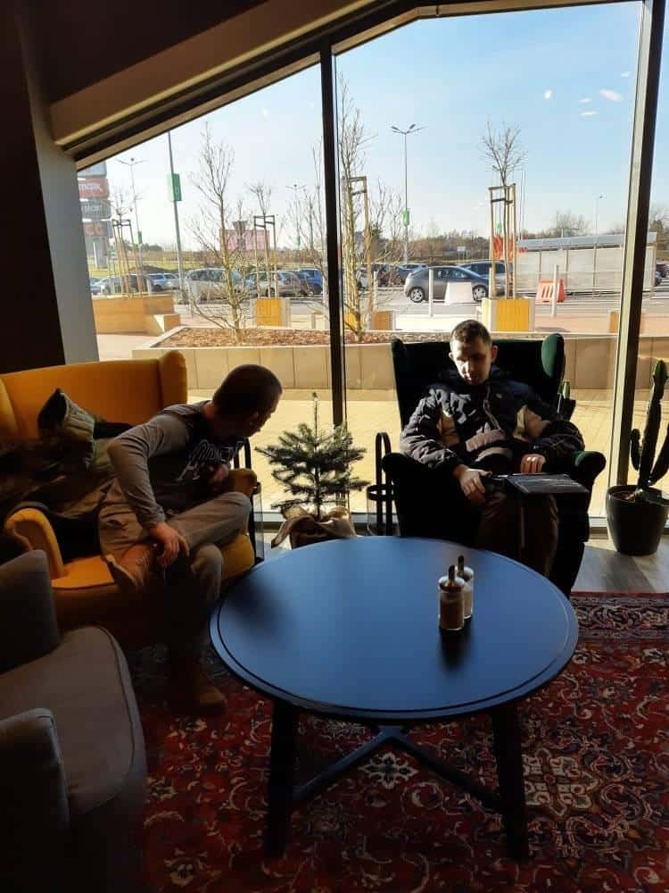 Dwie osoby siedzą w fotelach przy okrągłym stole przed dużym oknem, z małym psem między nimi i parkingiem widocznym na zewnątrz.