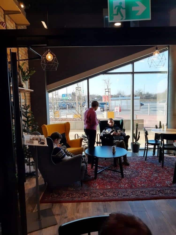 Trzy osoby siedzą i rozmawiają w fotelach wokół stolika w nowoczesnej kawiarni z dużymi oknami i naturalnym światłem.