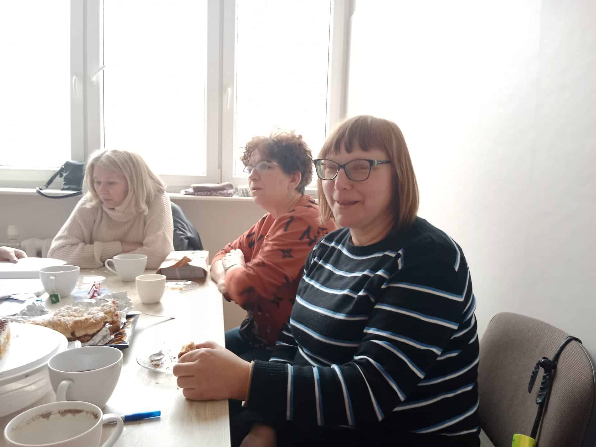 Trzy kobiety siedzą przy stole z filiżankami, talerzami i ciastem w jasno oświetlonym pokoju, zwrócone w stronę kamery.