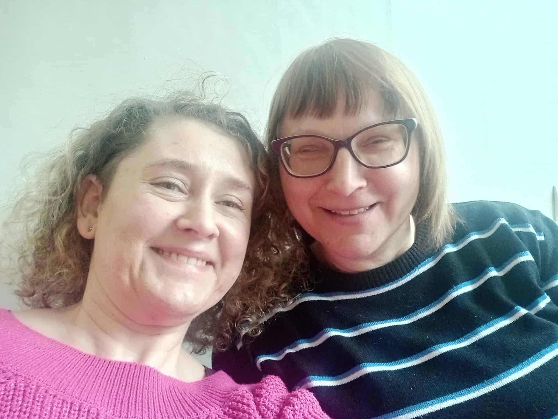 Dwie kobiety uśmiechające się do kamery, jedna ubrana w różowy sweter, a druga w czarny sweter w paski i okulary, na jasnym tle.