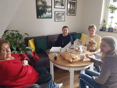 Cztery osoby siedzą przy okrągłym stole z napojami, jedzeniem i laptopem w salonie. Jedna osoba trzyma małego psa. Na ścianach znajdują się rośliny i oprawione zdjęcia.