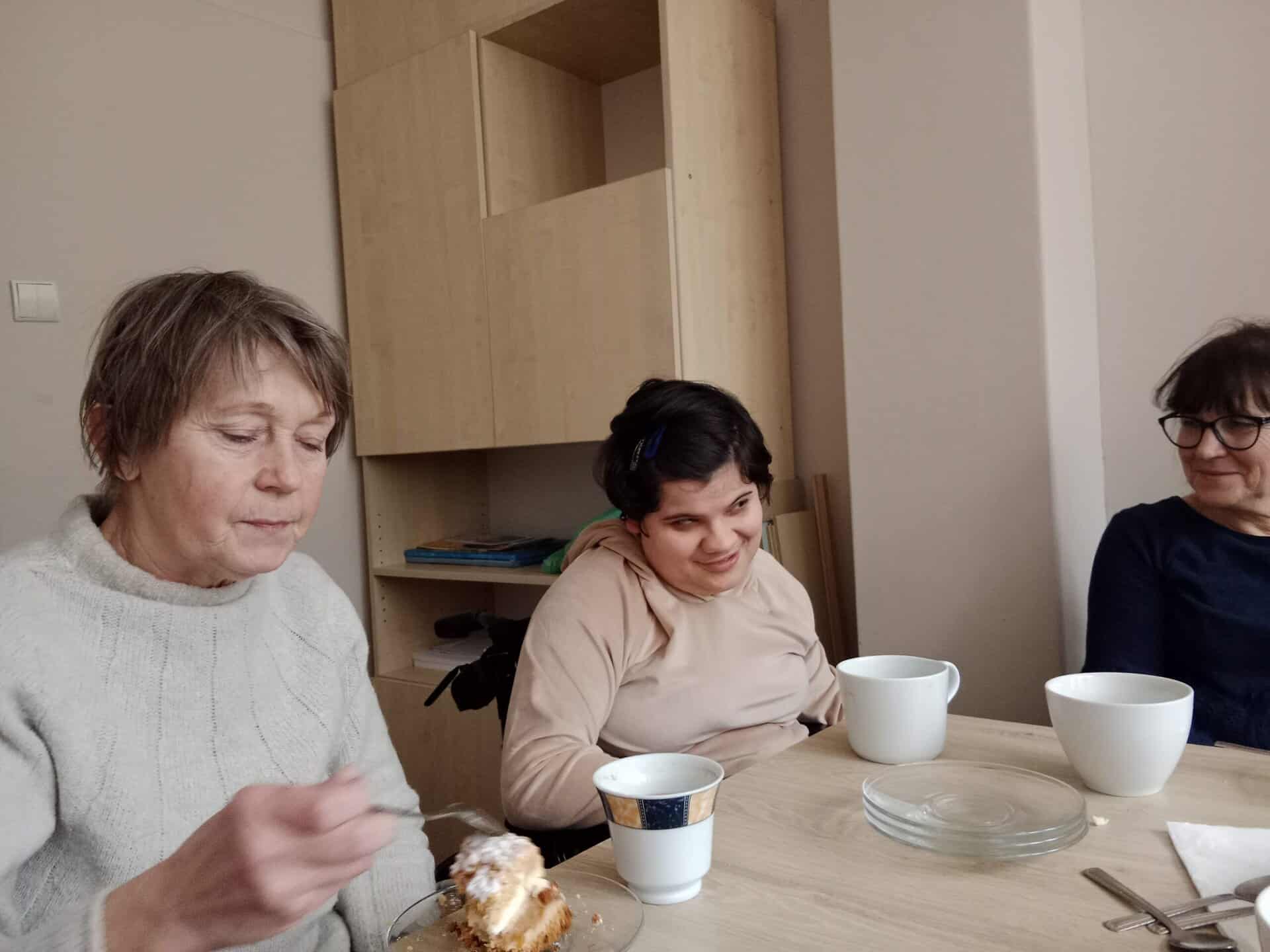 Trzy kobiety siedzą przy stole z filiżankami i deserem. Jedna z kobiet je ciasto, podczas gdy pozostałe siedzą w pobliżu, wszystkie wewnątrz w prosto urządzonym pokoju.