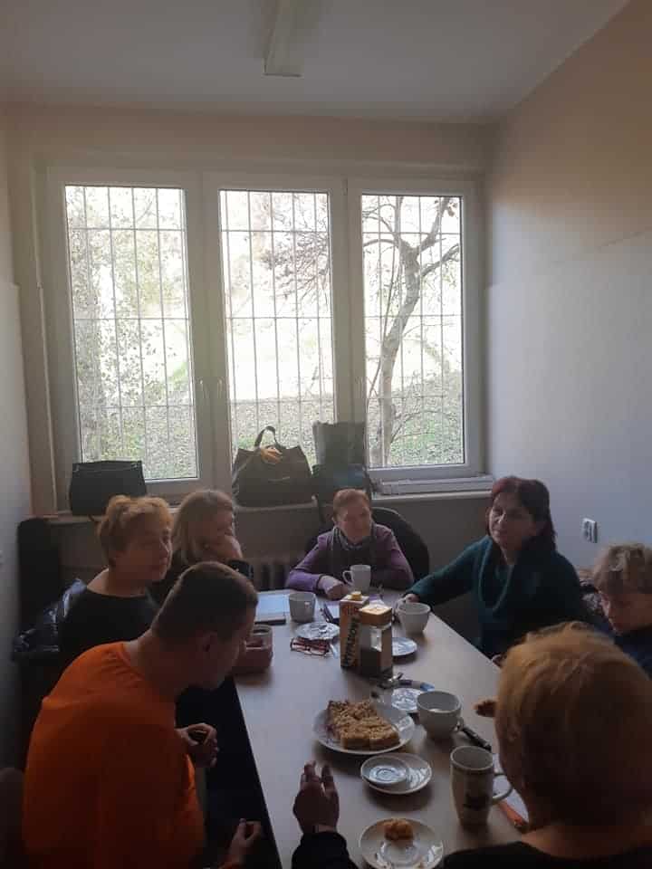 Siedem osób siedzi przy stole w małym pokoju, rozmawiając przy kawie i ciastkach, z oknem i torbami widocznymi w tle.