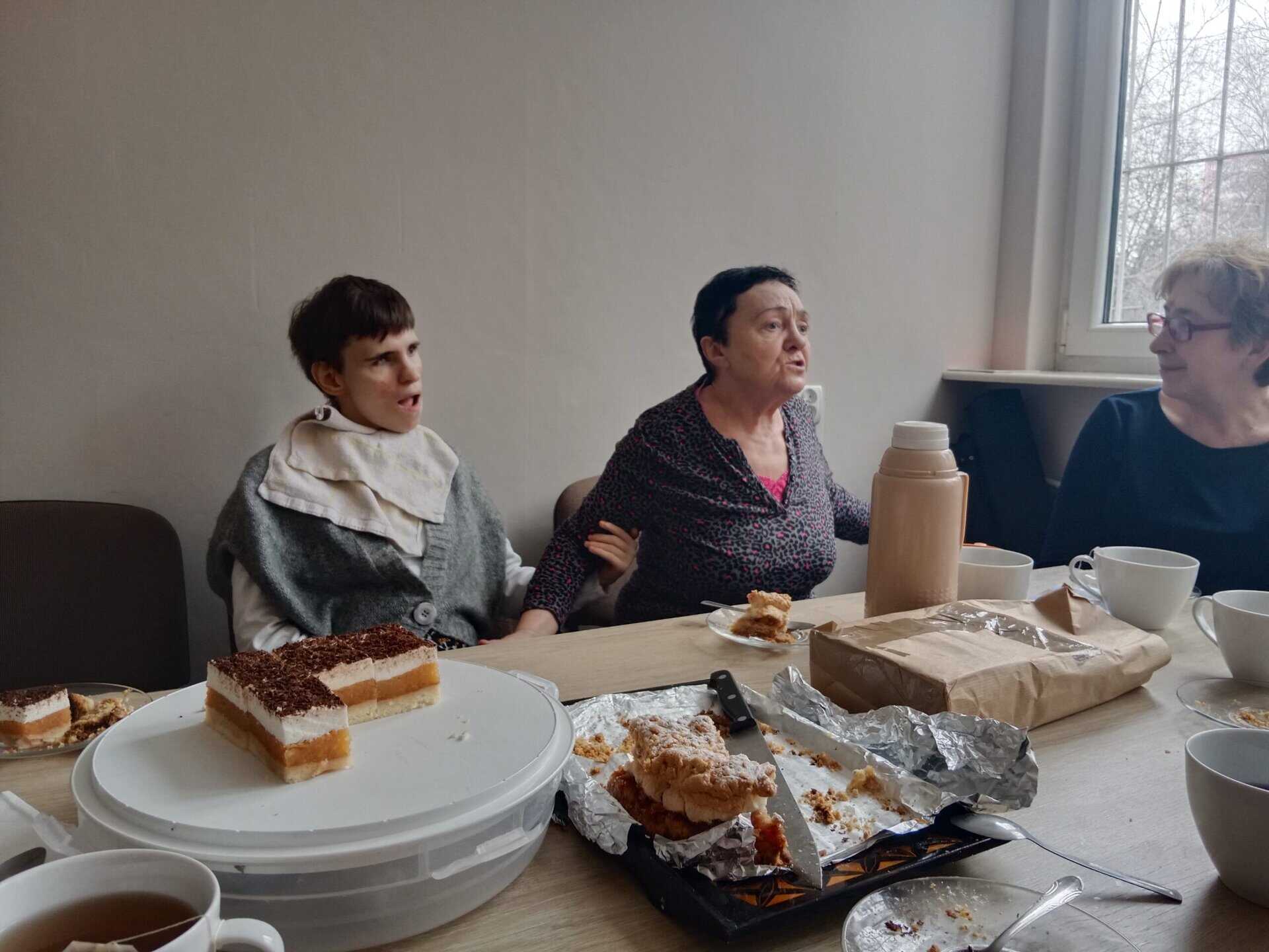 Trzy kobiety siedzą przy stole z ciastkami, filiżankami i termosem, pogrążone w rozmowie. Okno wpuszcza światło dzienne z prawej strony.