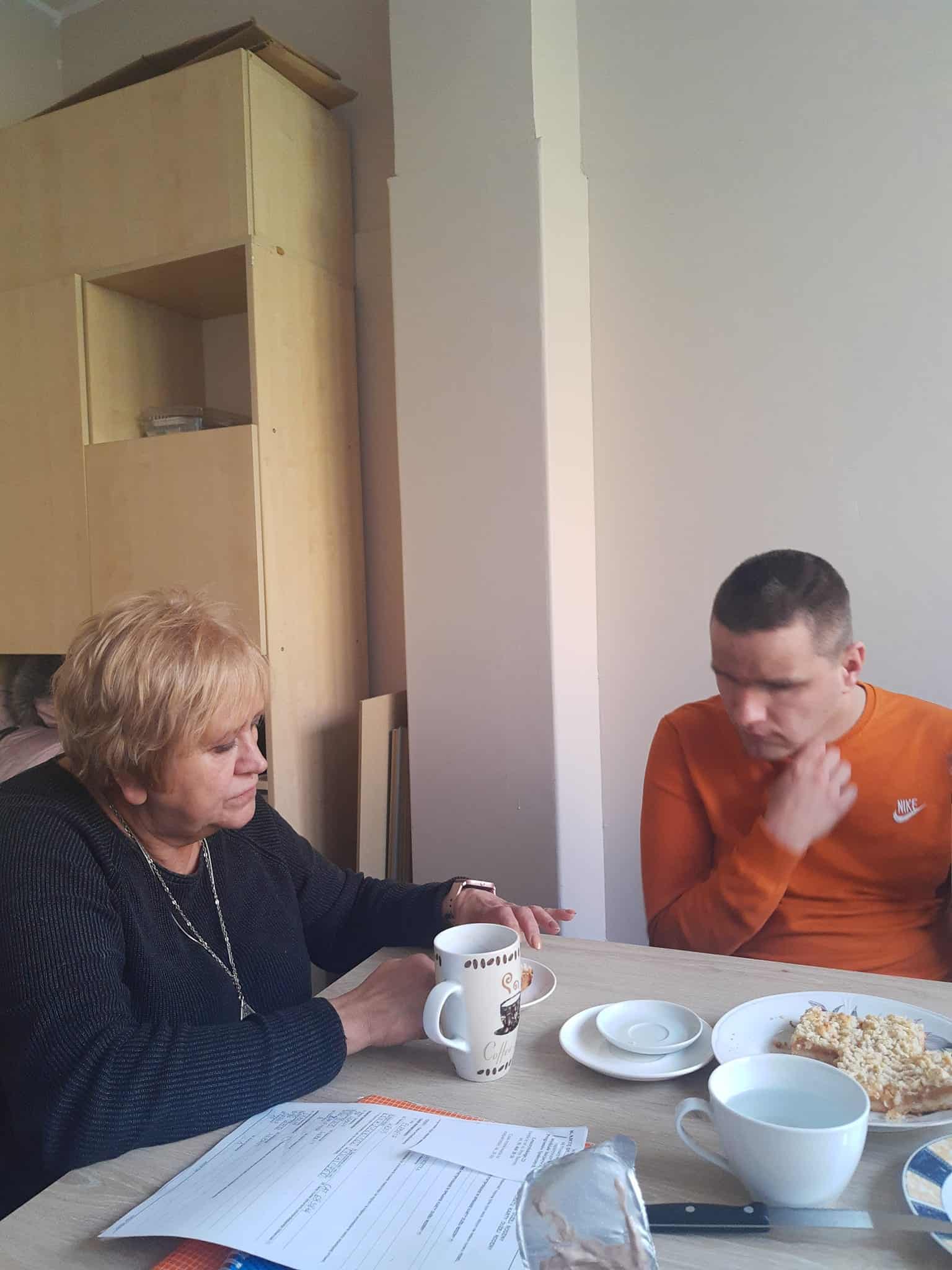 Dwie osoby siedzą przy stole z kubkami, talerzem jedzenia i dokumentami. Kobieta wydaje się mówić, podczas gdy mężczyzna słucha z ręką na brodzie.