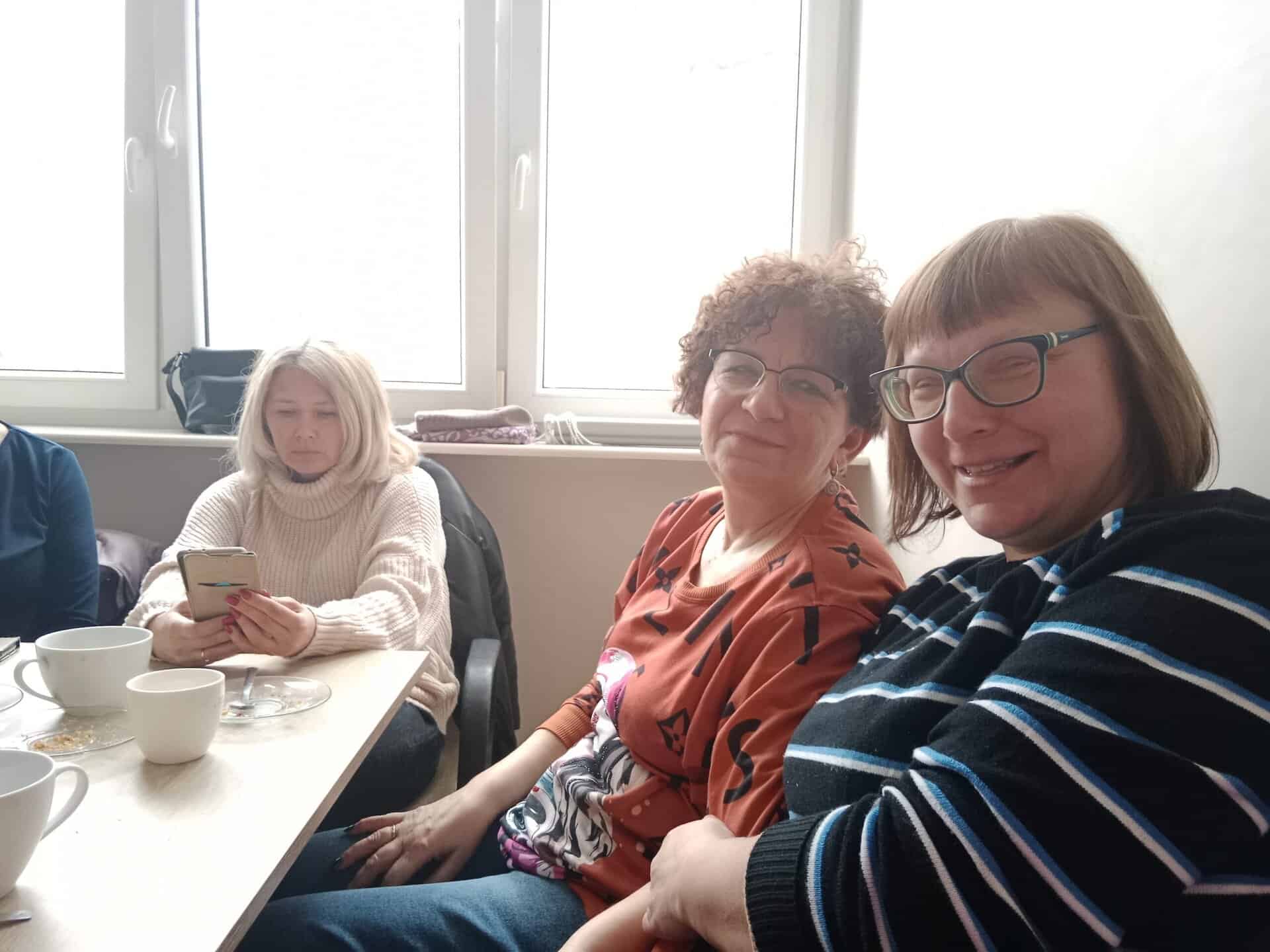 Trzy kobiety siedzą wokół stołu z filiżankami i talerzami; jedna używa telefonu, a dwie uśmiechają się do kamery w jasno oświetlonym pokoju z oknami.