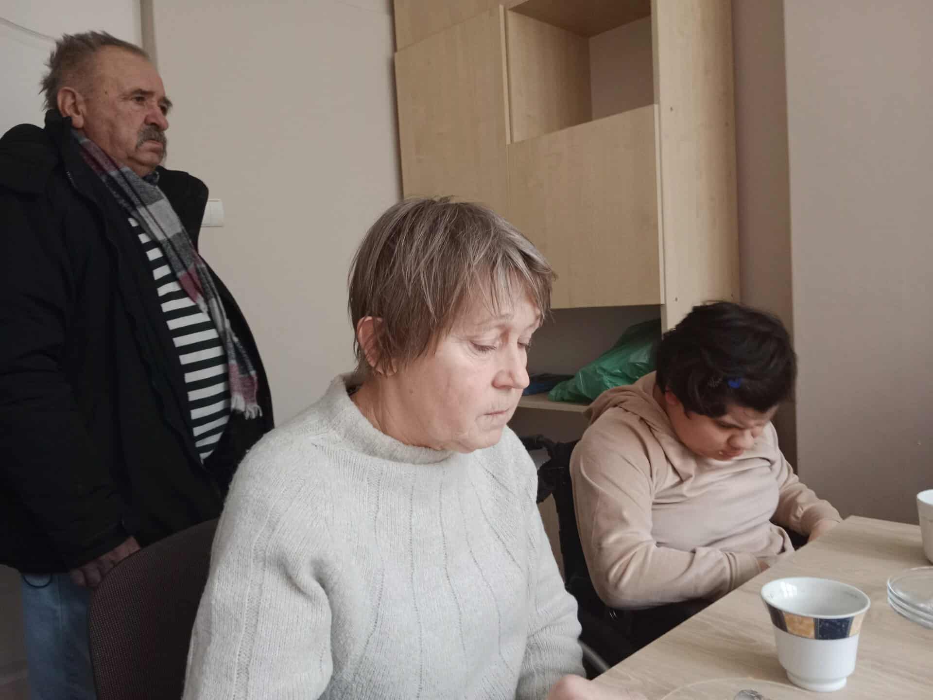 Trzy dorosłe osoby siedzą wokół stołu z filiżankami; dwie siedzą, a jedna stoi w tle. Atmosfera wydaje się spokojna i cicha.