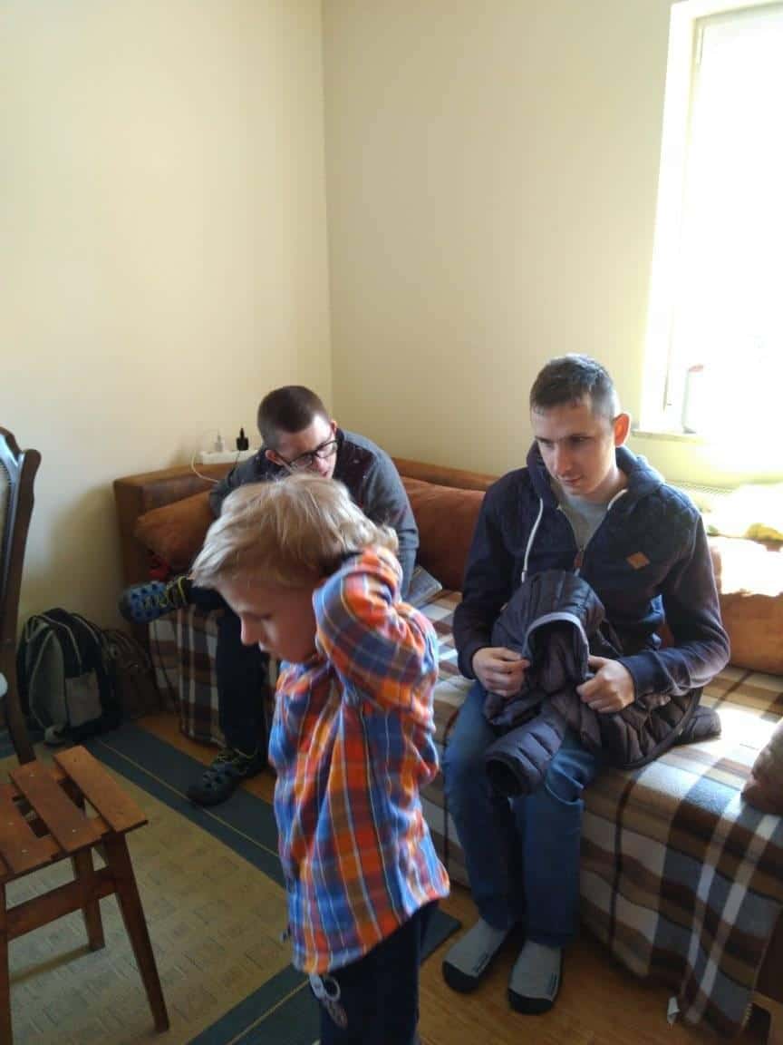 Trzy osoby znajdują się w pokoju; dwoje dorosłych siedzi na kanapie pokrytej kratą, a dziecko w kraciastej koszuli stoi na pierwszym planie, dopasowując rękawy.