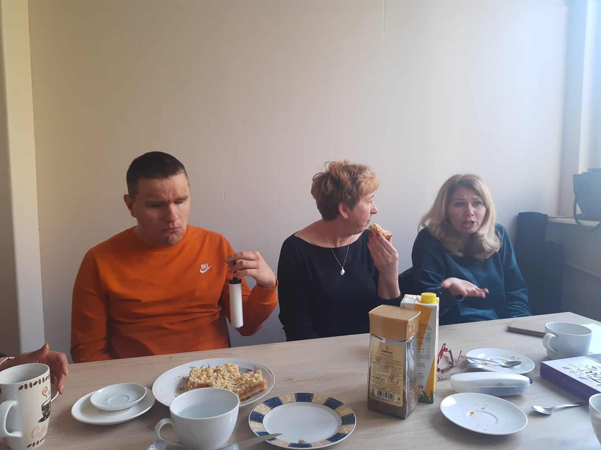 Trzy osoby siedzą przy stole z talerzami, kubkami i różnymi przedmiotami śniadaniowymi, jedząc i rozmawiając w naturalnie oświetlonym pokoju.