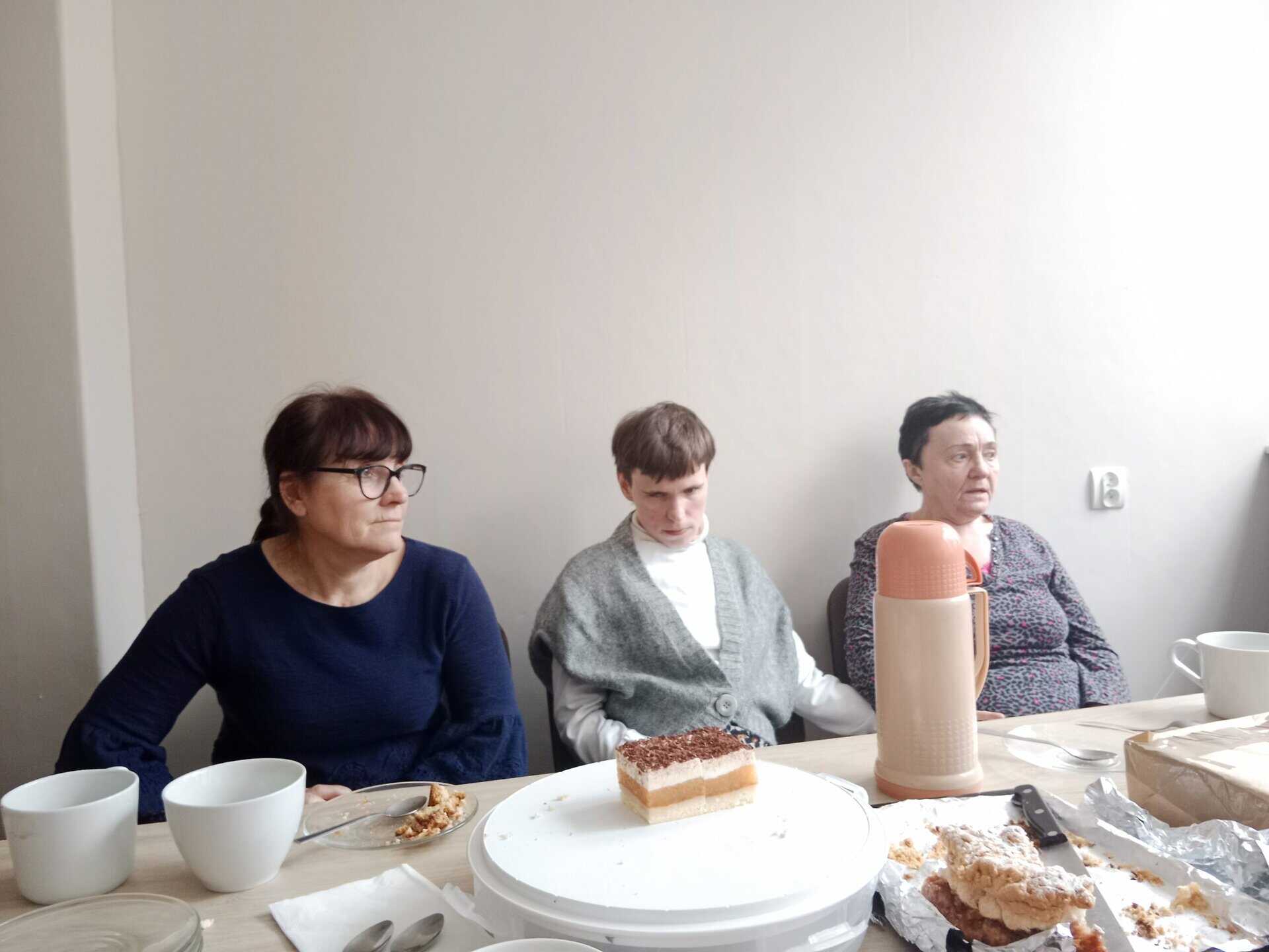Trzy kobiety siedzą przy stoliku z filiżankami, talerzami, ciastem i termosem przed sobą, na tle prostej, jasnej ściany.