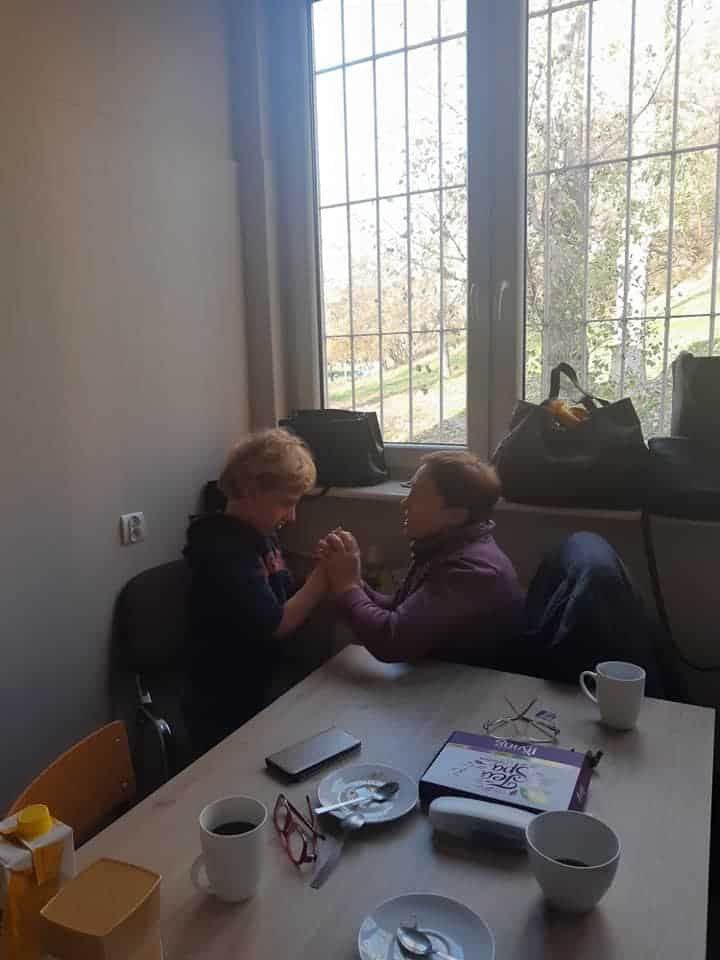 Dziecko i dorosły siłują się na ręce przy stole z kubkami, sokiem, telefonem i książką w jasnym pokoju z oknem i torbami na parapecie.