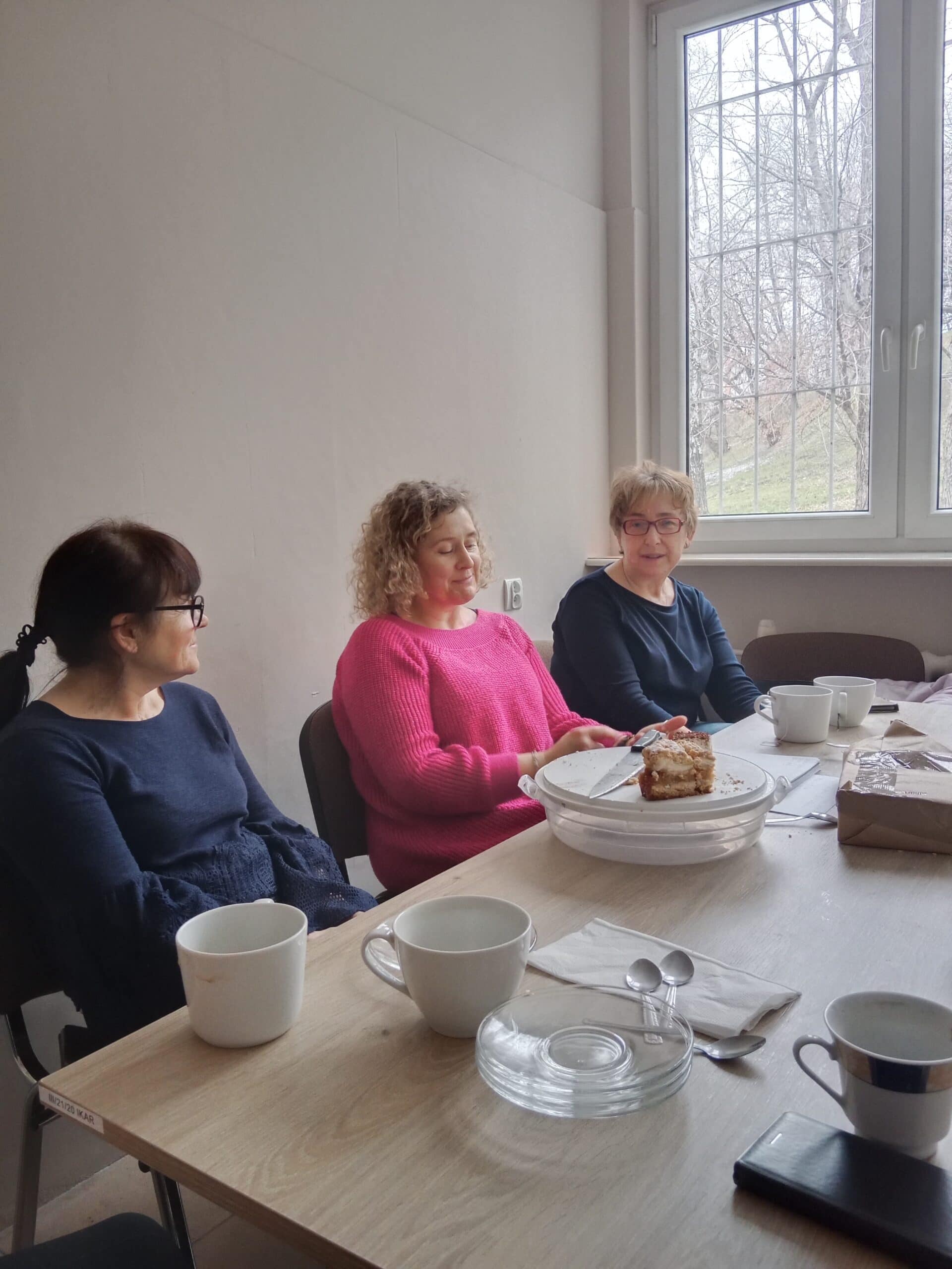 Trzy kobiety siedzą przy stole z pustymi filiżankami i talerzami, rozmawiając po zjedzeniu ciasta w jasnym pokoju z oknem ukazującym drzewa na zewnątrz.