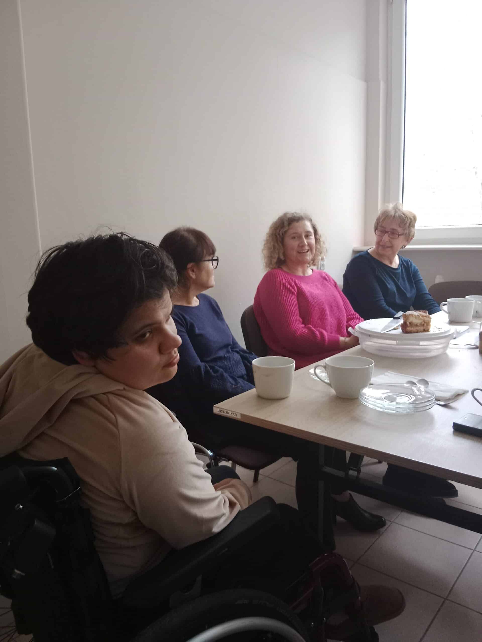 Cztery kobiety siedzą wokół stołu z filiżankami, talerzami i deserem. Jedna z kobiet na pierwszym planie siedzi na wózku inwalidzkim i patrzy w stronę kamery.