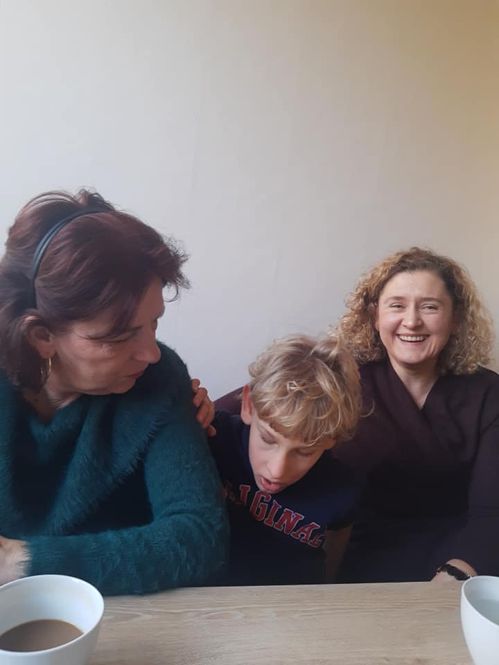 Trzy osoby siedzą przy stole z filiżankami. Kobieta w zieleni patrzy na chłopca o blond włosach w centrum, podczas gdy inna kobieta z kręconymi włosami uśmiecha się po prawej stronie.