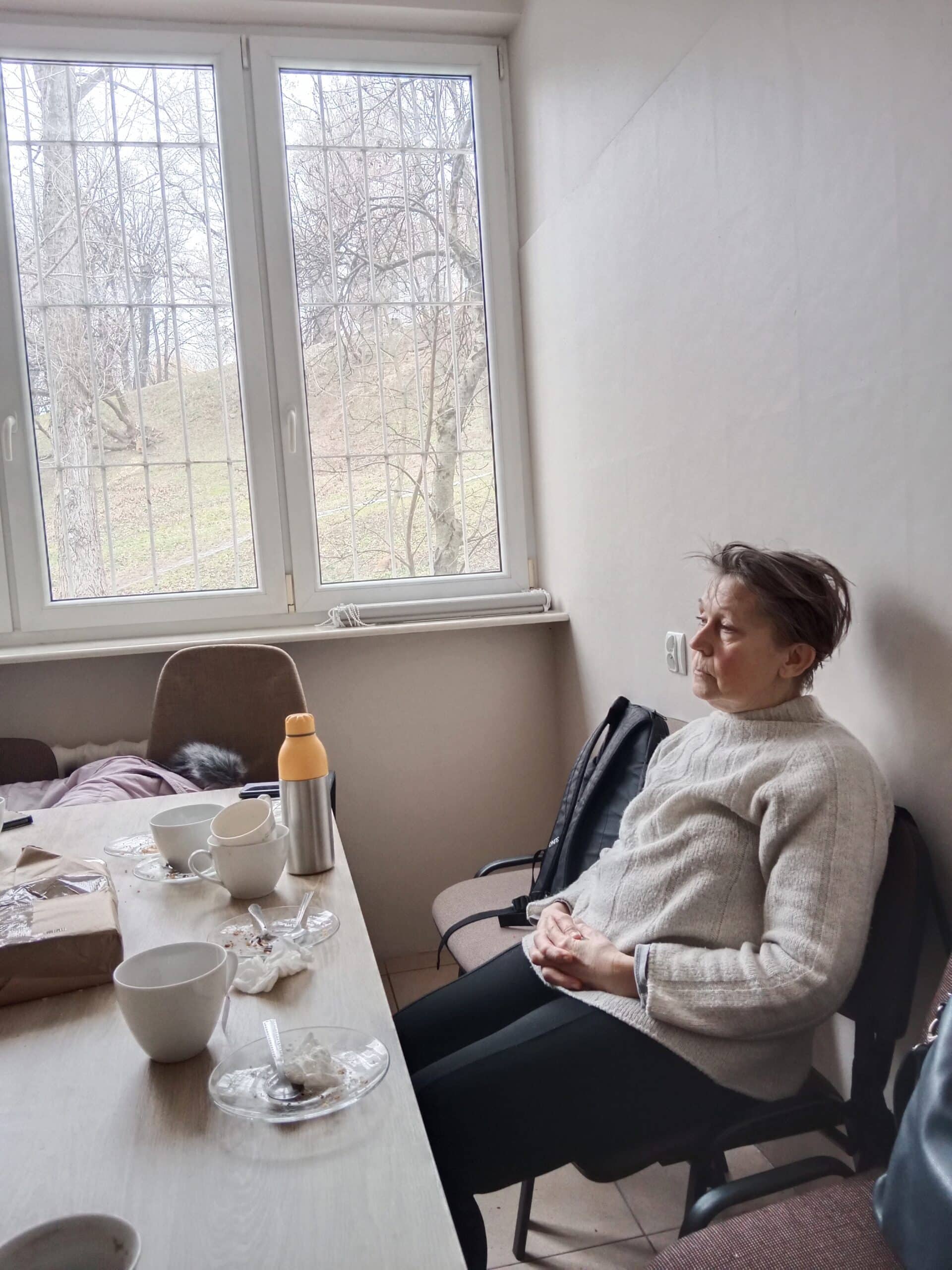 Osoba siedzi z zamkniętymi oczami i rękami złożonymi na kolanach w pokoju obok stołu ze zużytymi naczyniami i termosem; przez okno widać drzewa.