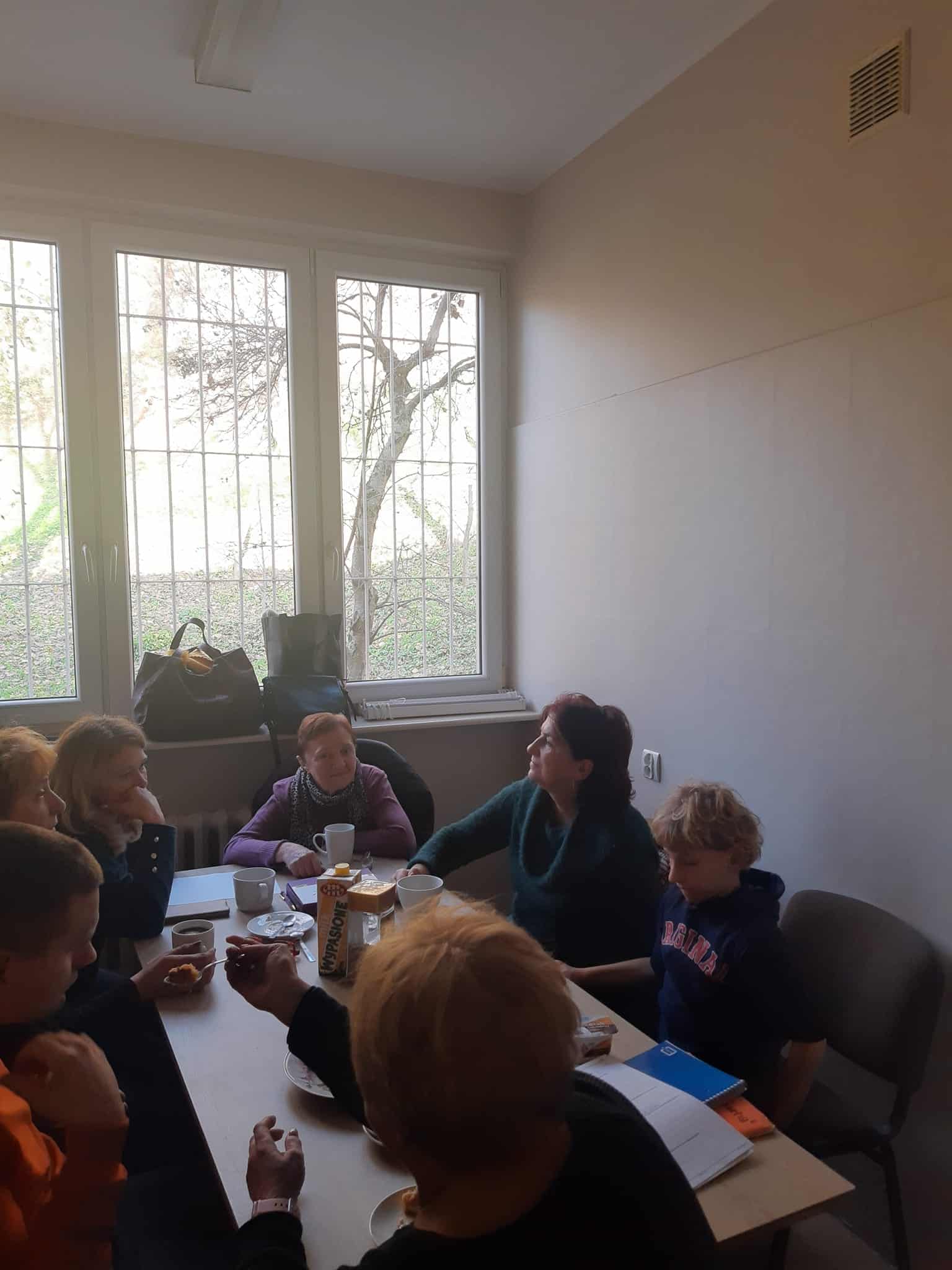 Grupa ludzi siedzi przy stole w małym pokoju i dyskutuje. Niektórzy mają napoje i przekąski, a przez zakratowane okno za nimi wpada światło dzienne.