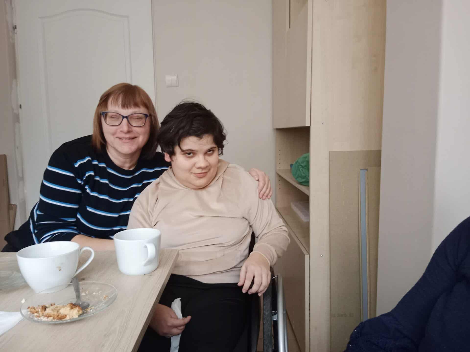 Dwie osoby siedzą razem przy stole z kubkami i talerzem jedzenia. Jedna osoba obejmuje drugą w geście wsparcia. Trzecia osoba jest częściowo widoczna po prawej stronie.