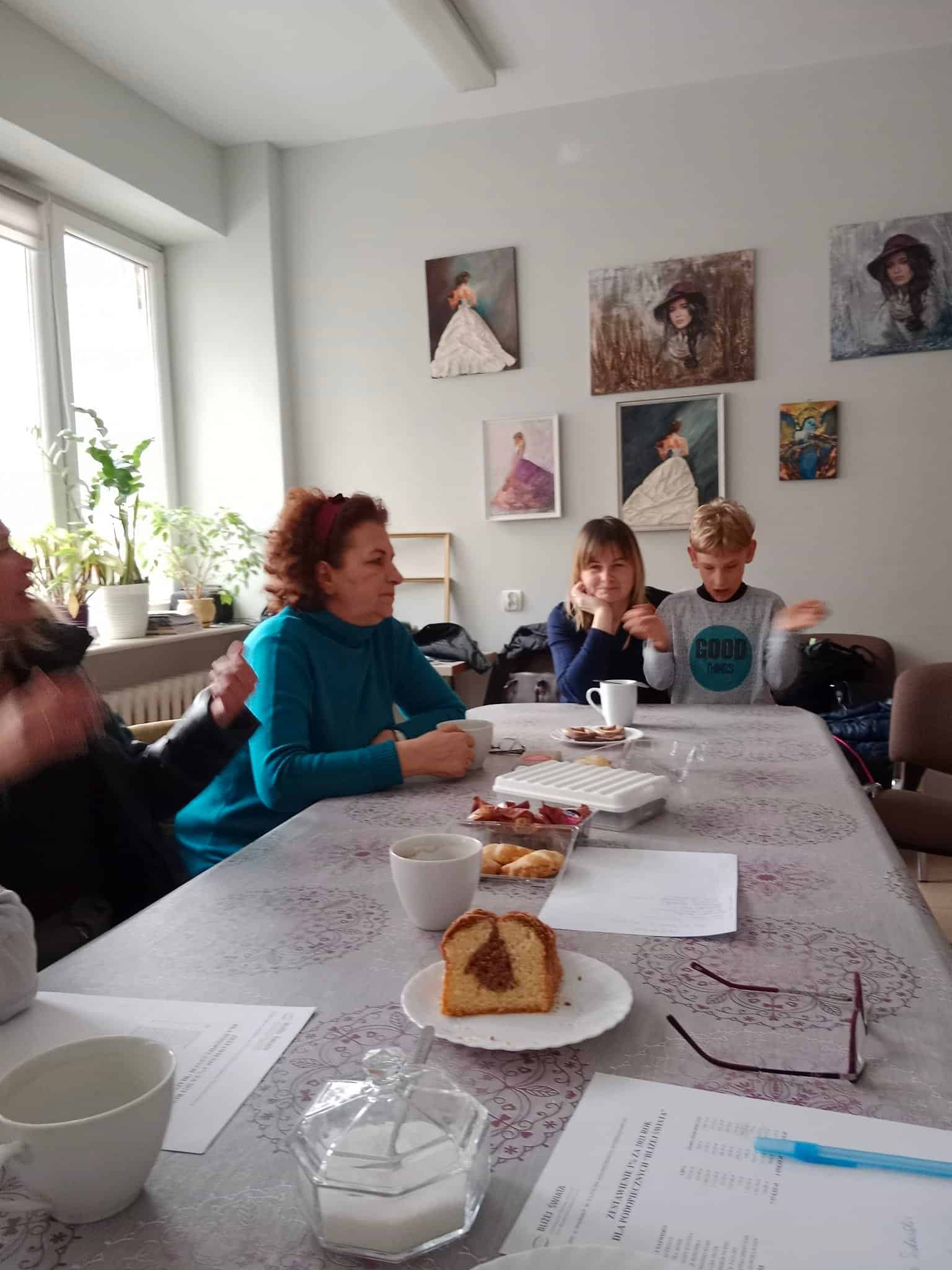 Cztery osoby siedzą przy stole z kawą, ciastkami i gazetami w pokoju ozdobionym portretami i roślinami doniczkowymi.