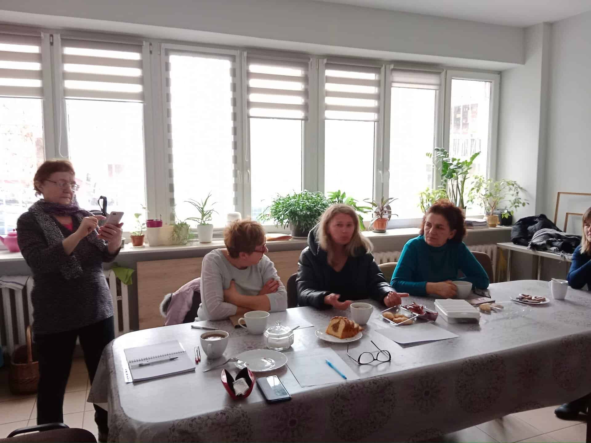 Pięć kobiet zgromadzonych wokół stołu z papierami, przekąskami i napojami; jedna z nich stoi i robi zdjęcie, podczas gdy pozostałe siedzą i rozmawiają.