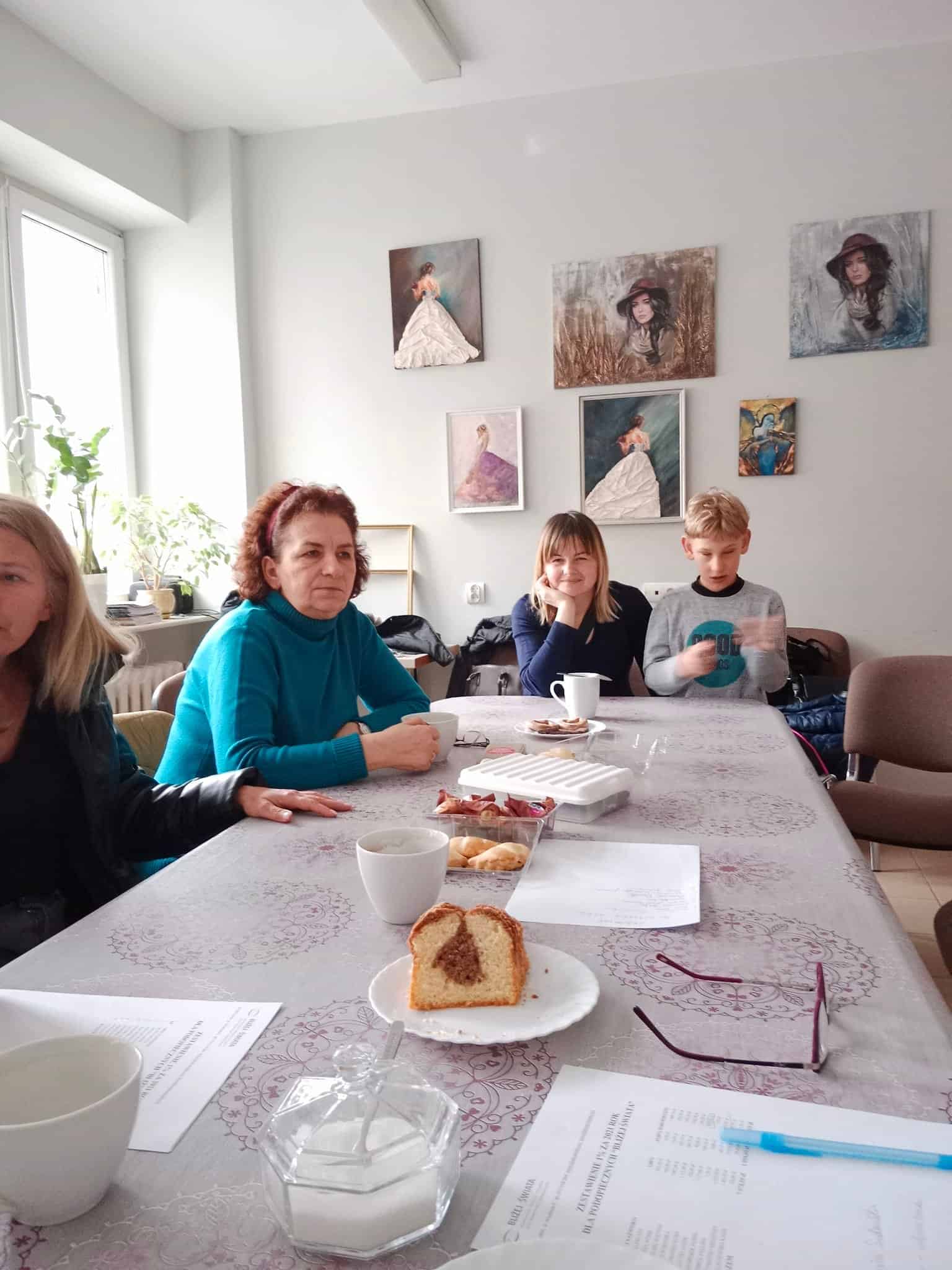 Ludzie siedzą wokół stołu z ciastkami, kubkami i gazetami, w pokoju ozdobionym portretami kobiet na ścianie.