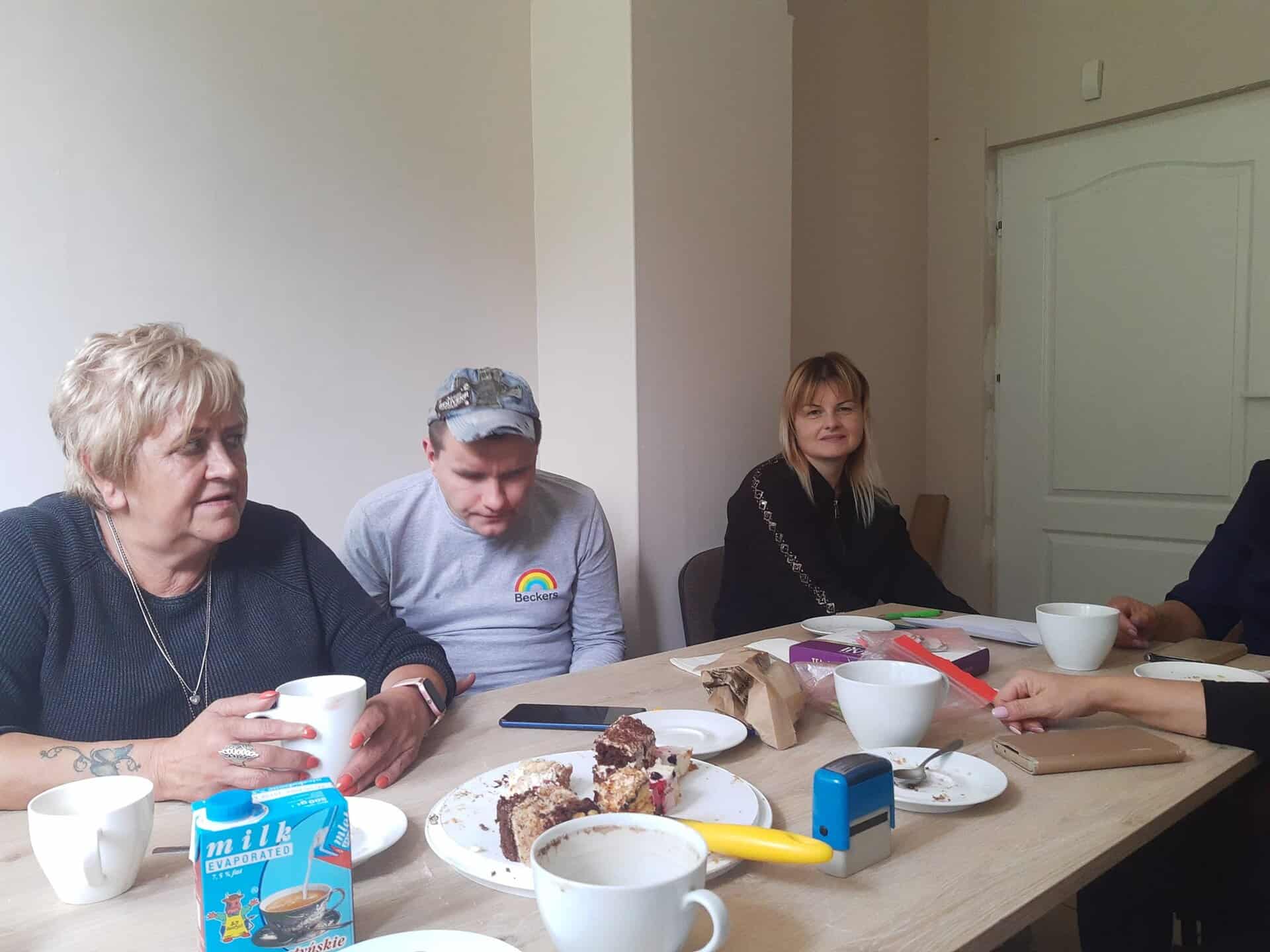 Cztery osoby siedzą przy stole z filiżankami kawy, ciastem, kartonem mleka i papierami, zaangażowane w dyskusję w zwykłym pokoju.