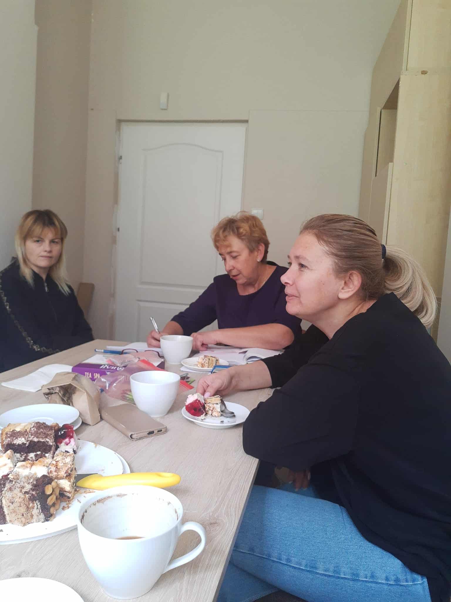 Trzy kobiety siedzą przy stole z kawą i ciastem, zaangażowane w rozmowę w prostym, jasnym pokoju.