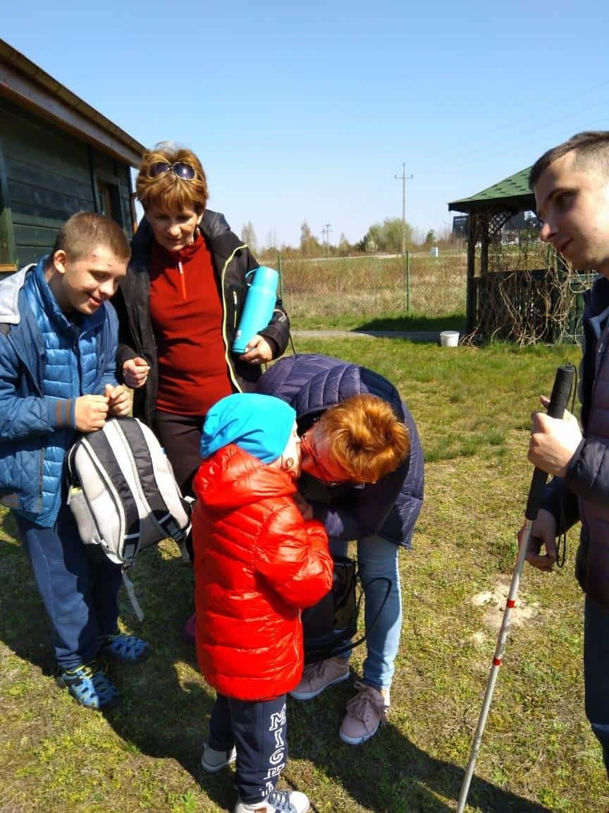 Pięć osób, w tym dwoje dorosłych i dziecko w czerwonej kurtce, zebranych na zewnątrz w pobliżu budynku; jedna osoba trzyma białą laskę.