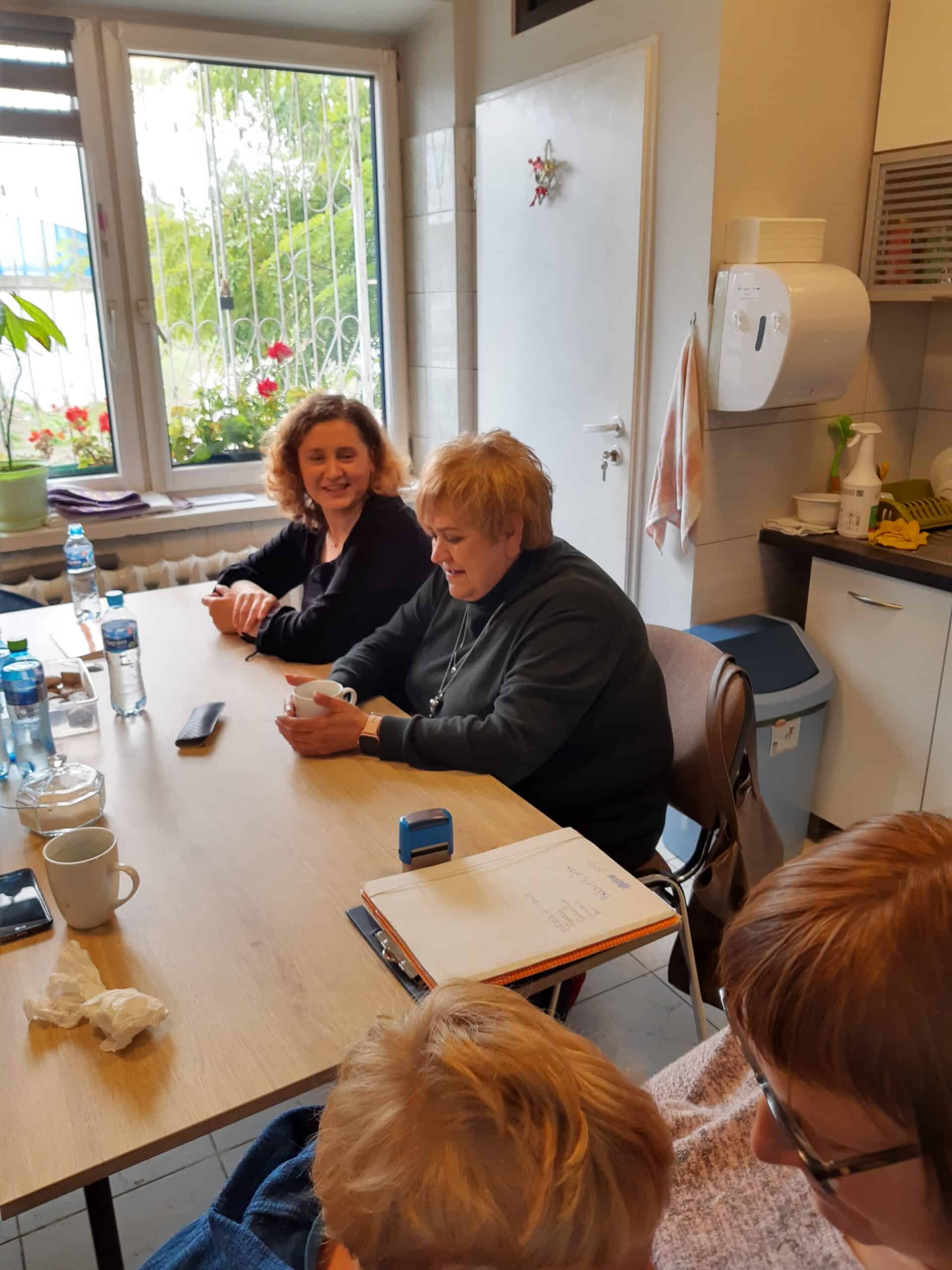 Cztery kobiety siedzą i rozmawiają przy stole w kuchni, mając przed sobą notatniki, kubki i butelki z wodą. W tle widać okno z roślinami.