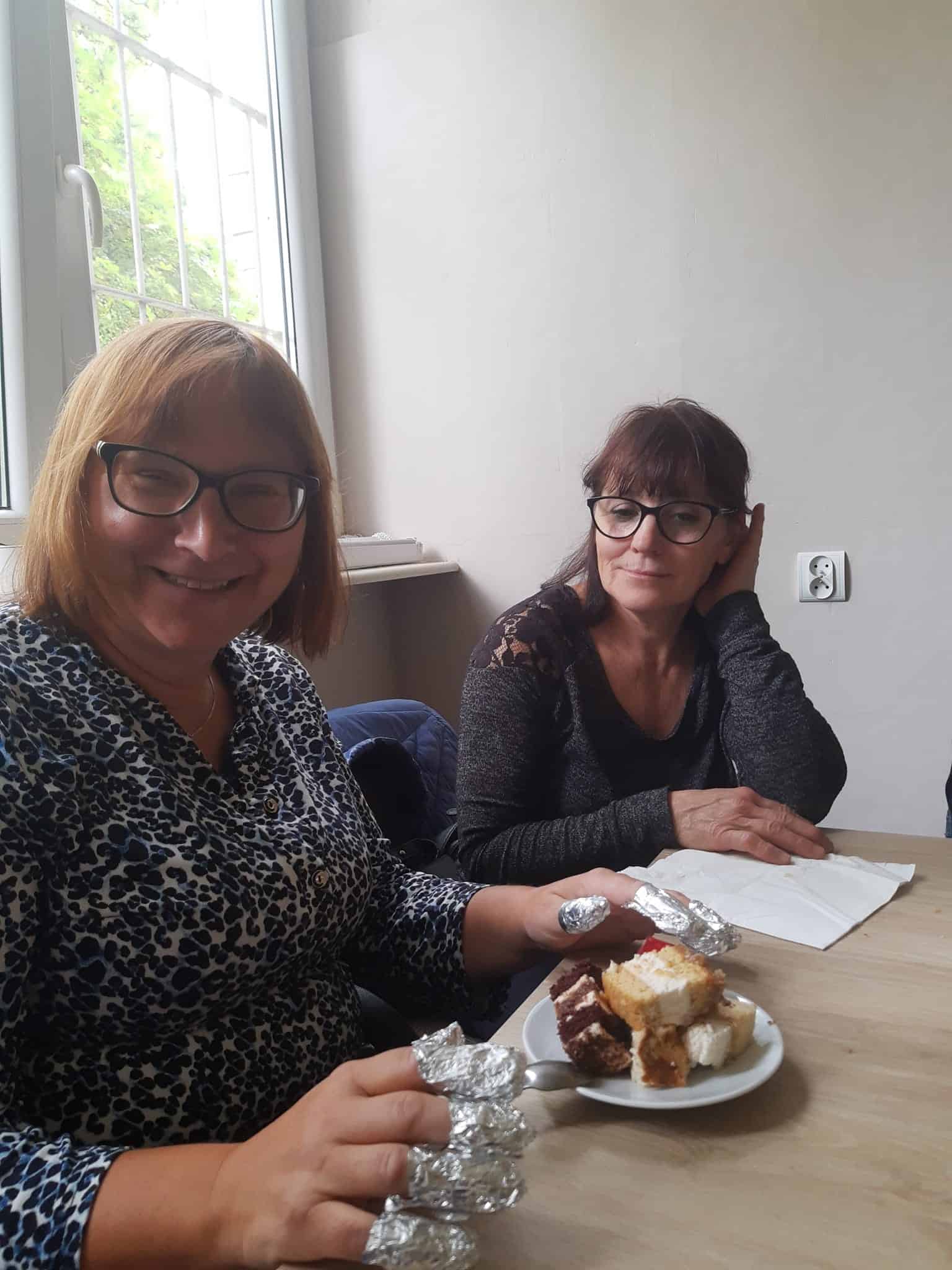 Dwie kobiety siedzą przy stole; jedna uśmiecha się i trzyma talerz z kawałkami ciasta zawiniętymi w folię, podczas gdy druga kobieta patrzy i pisze w notatniku.