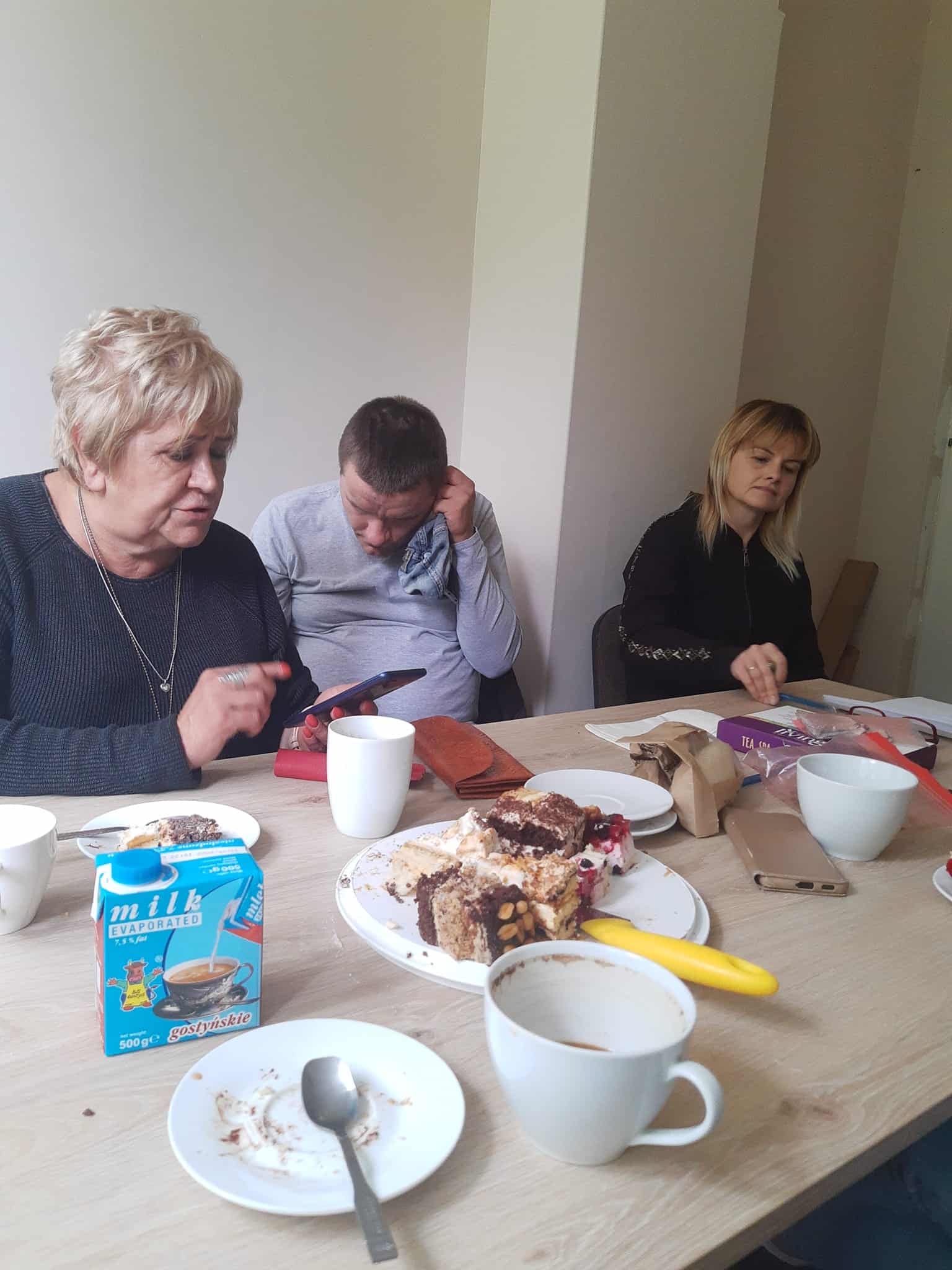 Trzy osoby siedzą przy stole z częściowo zjedzonym ciastem, kubkami, łyżeczkami i kartonem po napoju mlecznym. Jedna osoba korzysta z telefonu, a druga wyciera twarz serwetką.