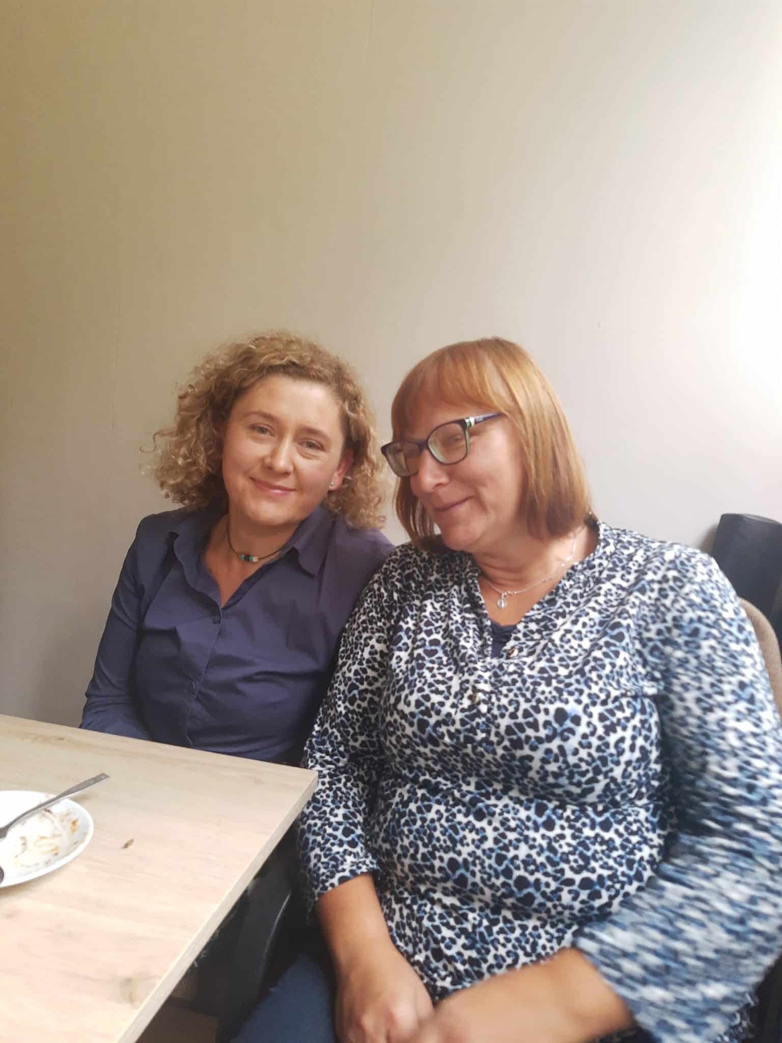 Dwie kobiety siedzą obok siebie przy stole w pomieszczeniu. Jedna z nich ma na sobie ciemnoniebieską koszulę, a druga niebiesko-biały wzorzysty top i okulary. Na stole stoi pusty talerz.