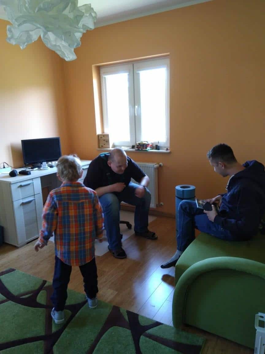 Trzy osoby znajdują się w jasno oświetlonym pokoju o pomarańczowych ścianach; dziecko stoi naprzeciwko dwóch dorosłych, z których jeden siedzi przy biurku, a drugi siedzi na zielonej kanapie i trzyma jakiś przedmiot.