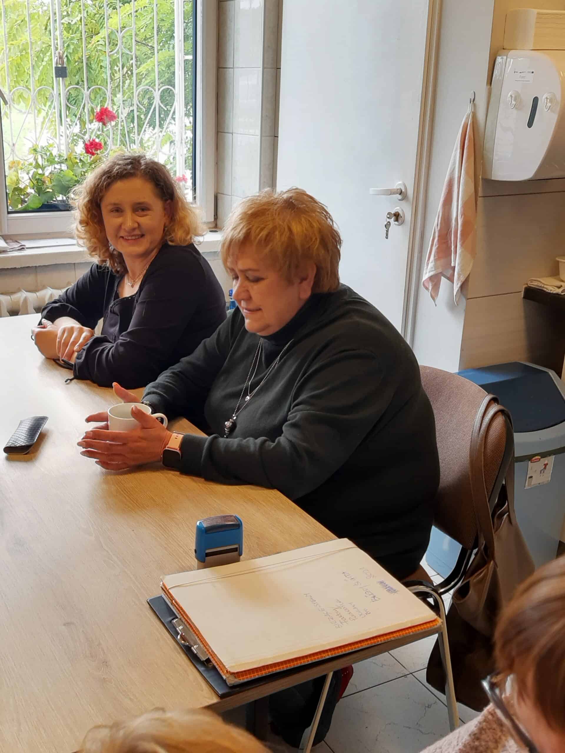 Dwie kobiety siedzą przy stole w pokoju, jedna mówi i gestykuluje rękami. Na stole znajduje się teczka z dokumentami, niebieska pieczęć i pilot.