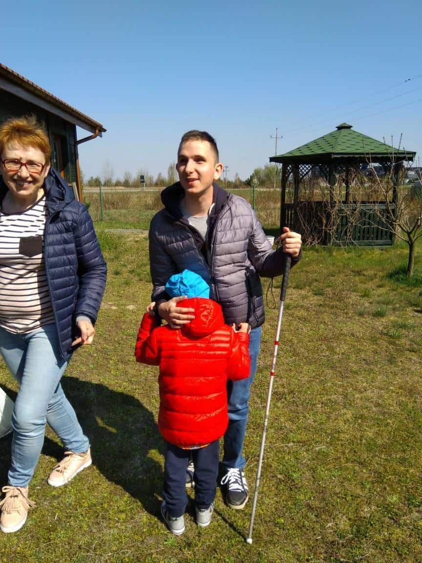 Trzy osoby stoją na świeżym powietrzu na trawie: uśmiechnięta kobieta po lewej stronie, mężczyzna trzymający białą laskę pośrodku i dziecko w czerwonej kurtce odwrócone od kamery.