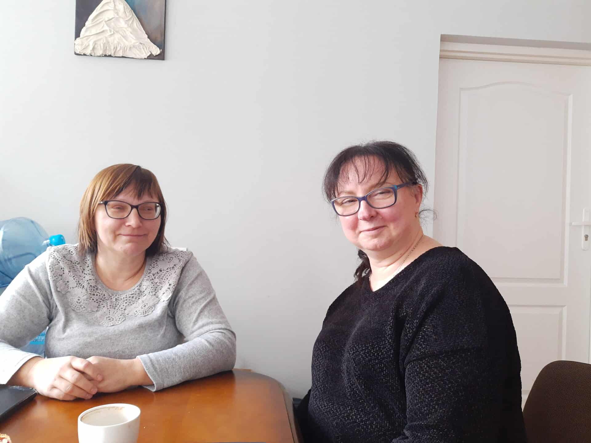 Dwie kobiety w okularach siedzą przy drewnianym stole z filiżanką kawy przed sobą, w prostym pokoju z białymi ścianami i zamkniętymi drzwiami.