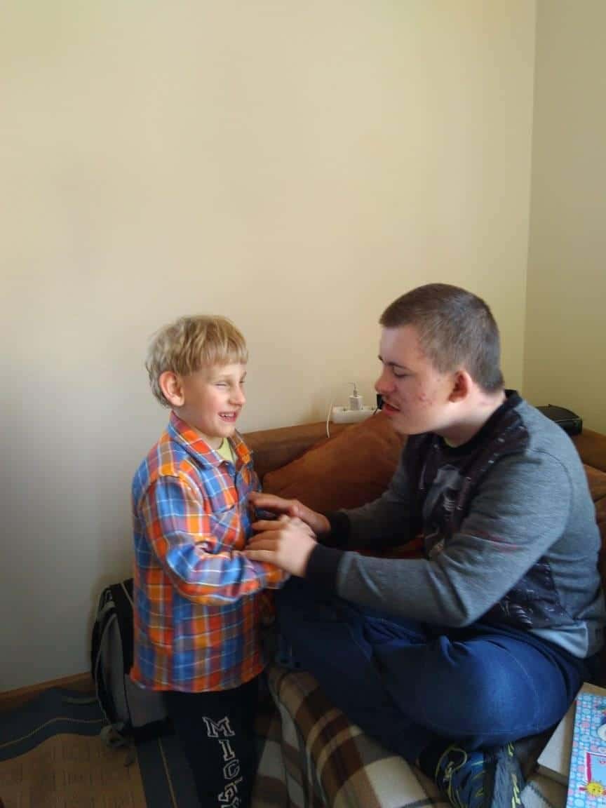 Młody chłopiec i starszy chłopiec z zespołem Downa siedzą na kanapie, uśmiechając się do siebie i trzymając się za ręce w jasno oświetlonym pokoju.