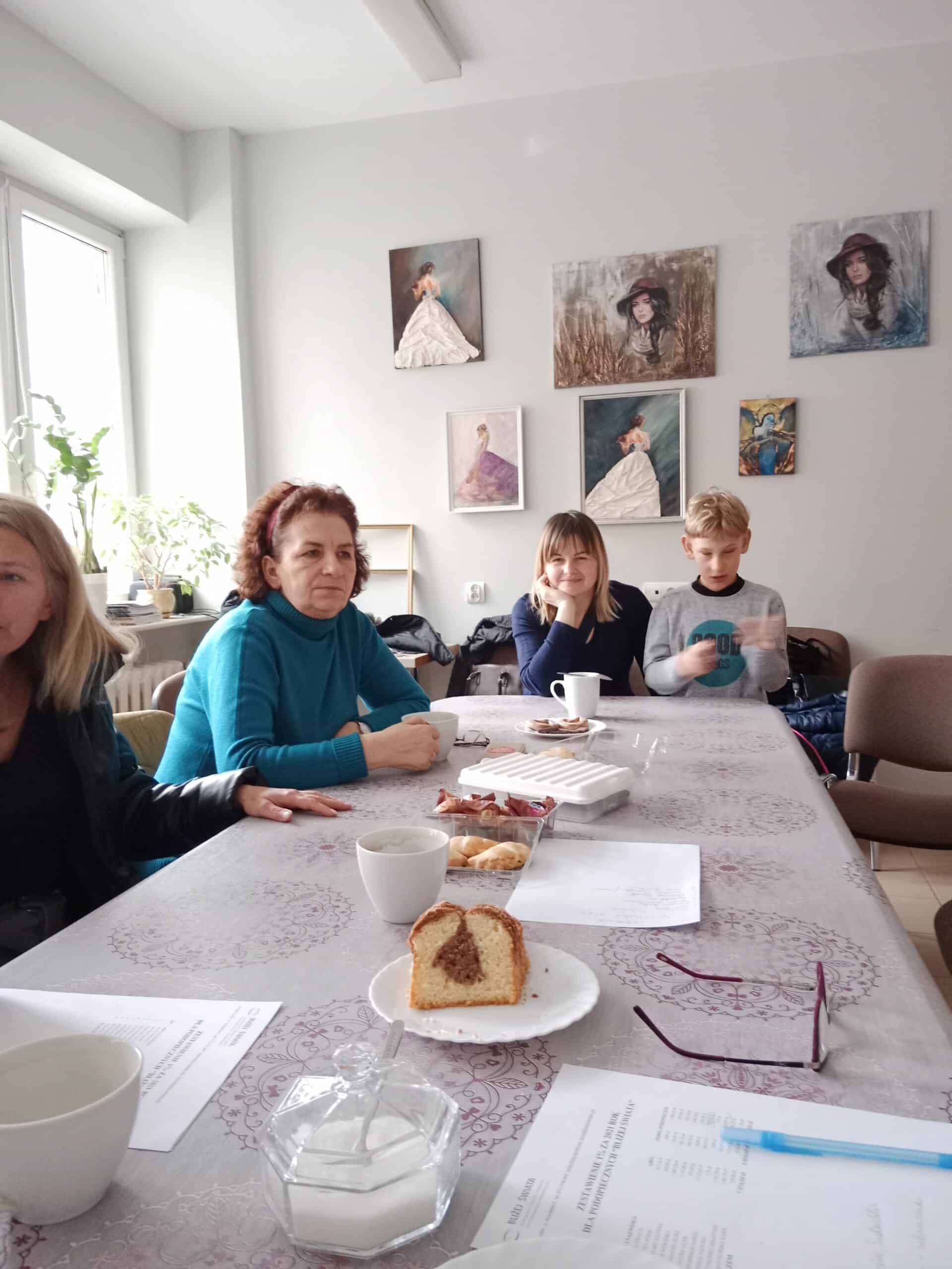 Ludzie siedzą wokół stołu z przekąskami, napojami i gazetami w jasnym pokoju ozdobionym obrazami kobiet i dziewcząt w sukienkach.