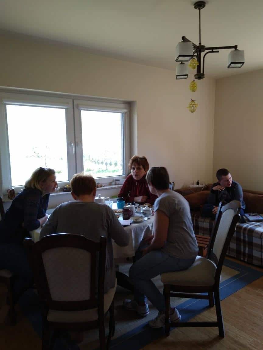 Pięć osób zebrało się w oświetlonym słońcem pokoju, cztery siedzą przy stole z jedzeniem i napojami, a jedna siedzi na kanapie w tle.