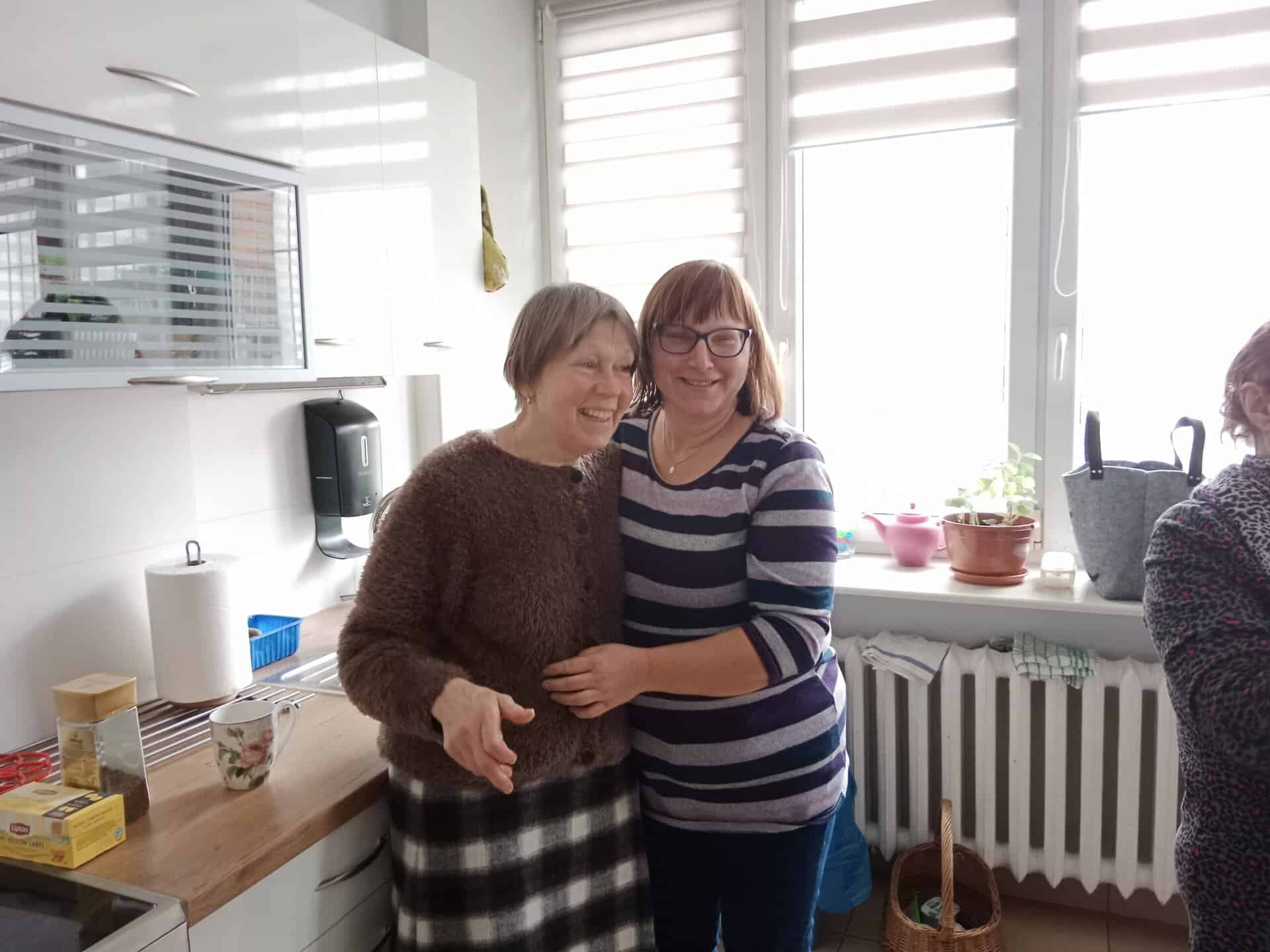 Dwie kobiety stoją uśmiechnięte i obejmujące się w jasnej kuchni obok lady z różnymi przedmiotami, z inną osobą częściowo widoczną po prawej stronie.