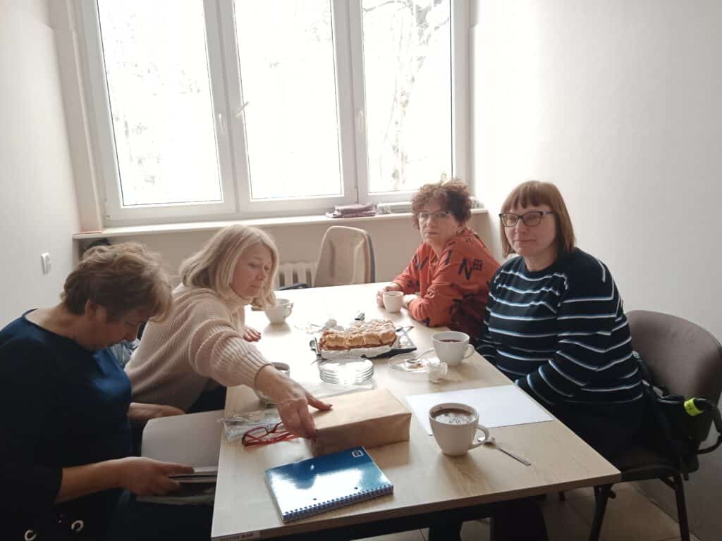 Cztery kobiety siedzą przy stole z kawą, gazetami i ciastem w jasno oświetlonym pokoju z dużym oknem.